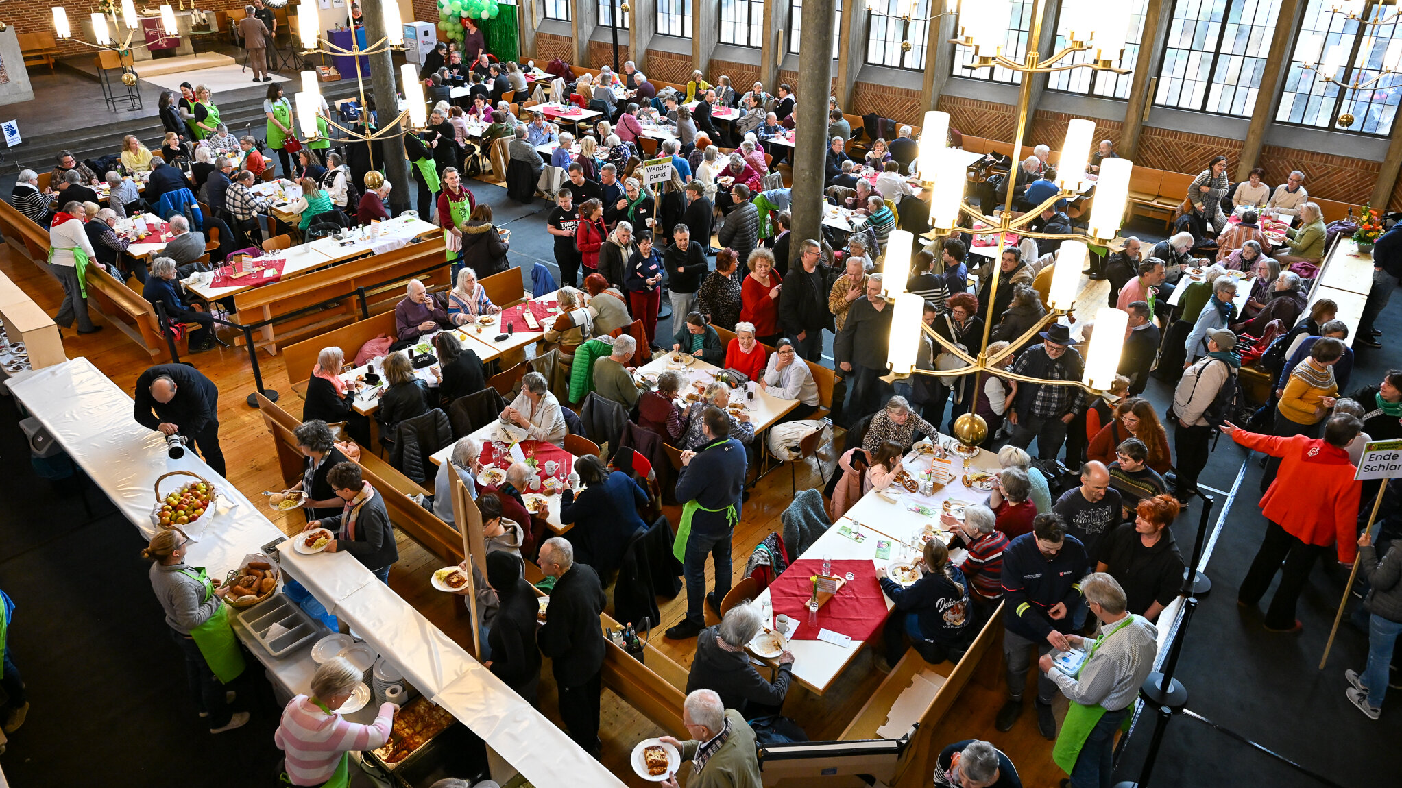 Das Foto zeigt den Innenraum einer Kirche. An der Seite sind Tische aneinander gereiht, an denen Essen von Menschen, die grüne Schürzen trage, ausgegeben wird. An vielen anderen Tische, sitzen Menschen und essen.