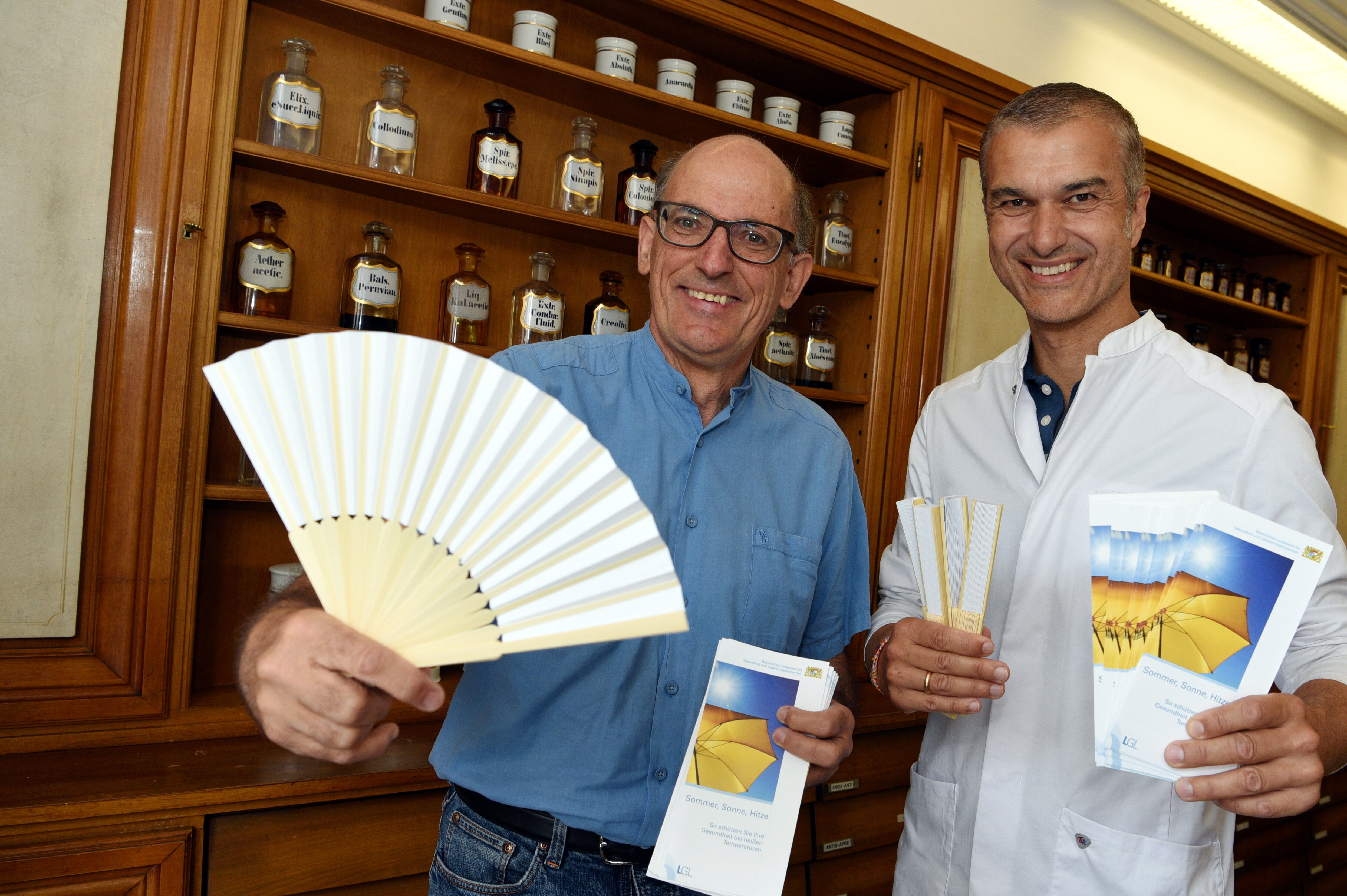 In einer Apotheke steht Umweltreferent Reiner Erben mit einem Fächer in der Hand. Neben ihm steht ein Apotheker mit weißem Kittel, er hält einige Flyer in der Hand.
