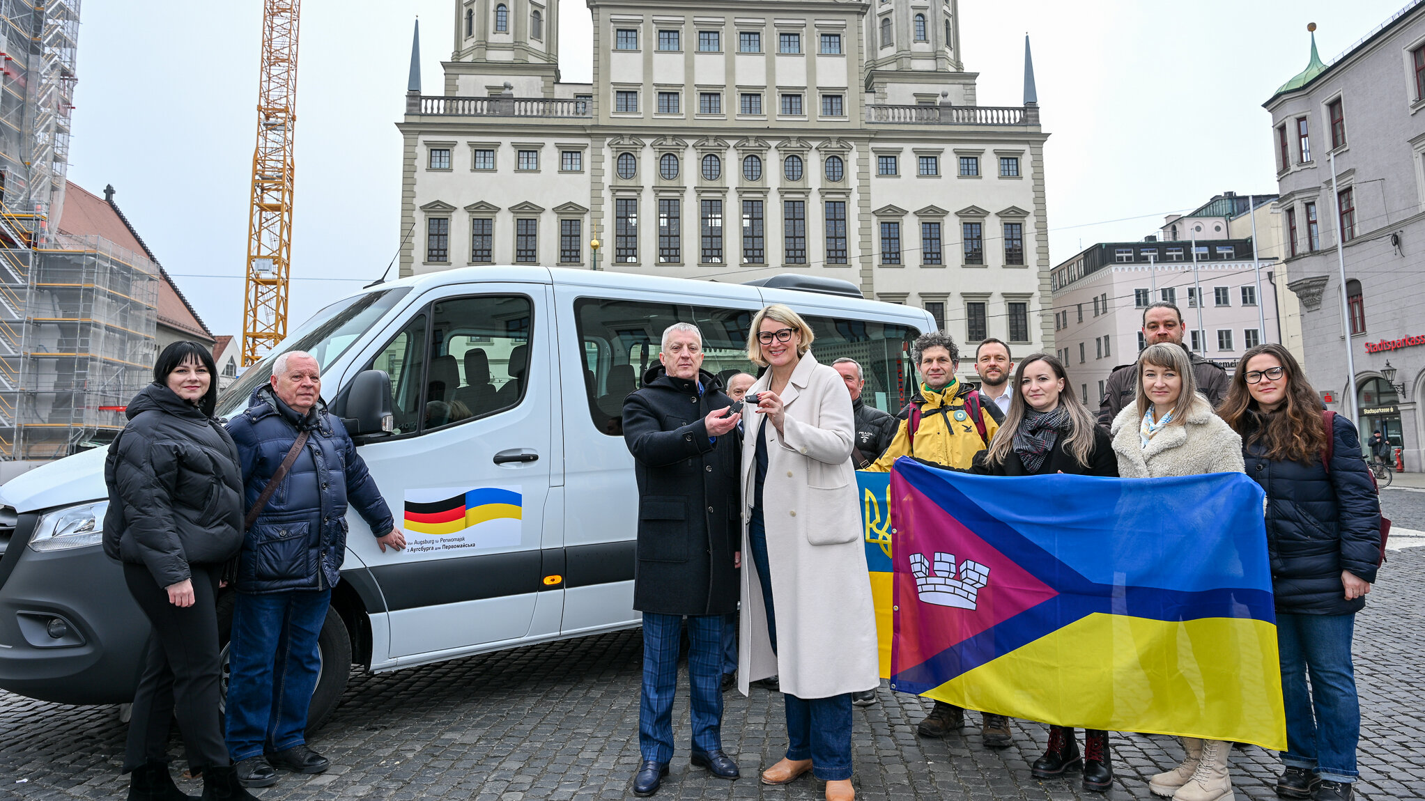 Das Foto zeigt eine Gruppe von Menschen auf dem Rathausplatz. In der Mitte steht Eva Weber und der Bürgermeister von Perwomajsk bei der Schlüsselübergabe. Im Hintergrund ist das neue Fahrzeug zu sehen. Darauf ist eine deutsche und eine ukrainische Flagge abgebildet