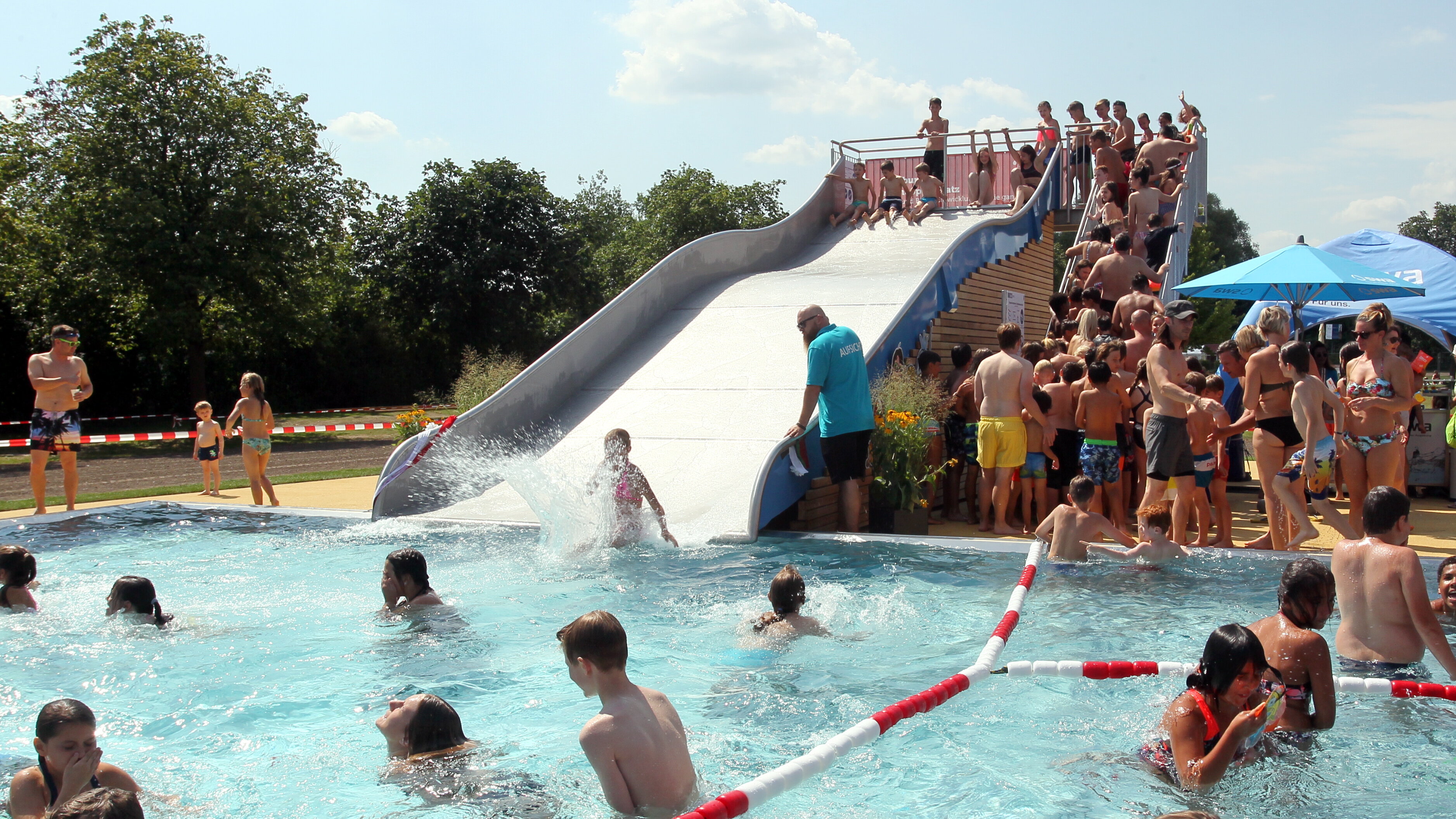 Das Foto zeigt ein Schwimmbecken in einem Freibad mit einer Rutsche. An der Rutsche stehen viele Kinder an.