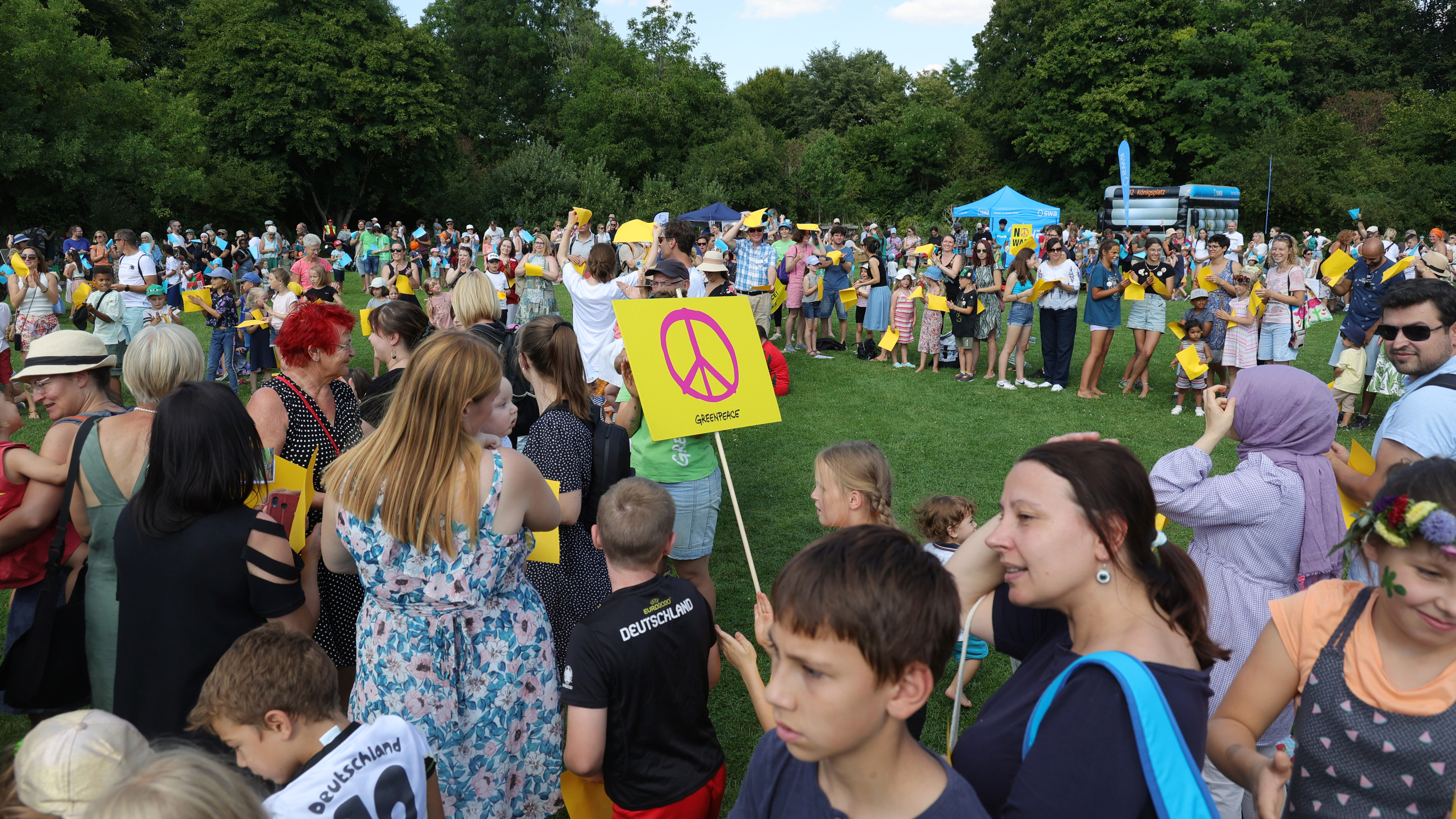 Viele Menschen unterschiedlichen Alters stehen auf einer von Bäumen umsäumten Rasenfläche, ein Kind hält ein Pappschild mit einem Peace-zeichen hoch