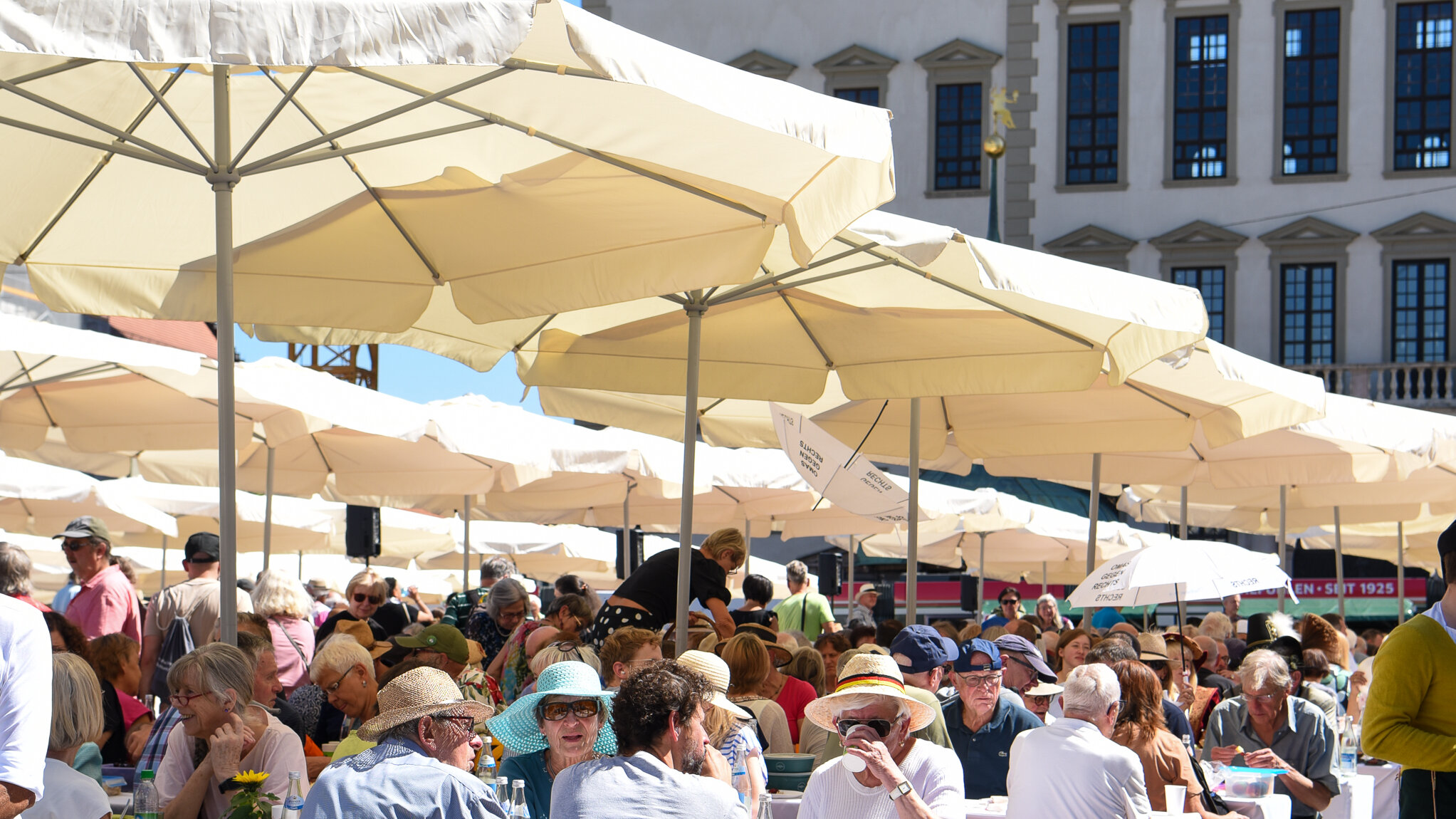 Viele Menschen sitzen unter hellen Sonnenschirmen beim Essen und Trinken.