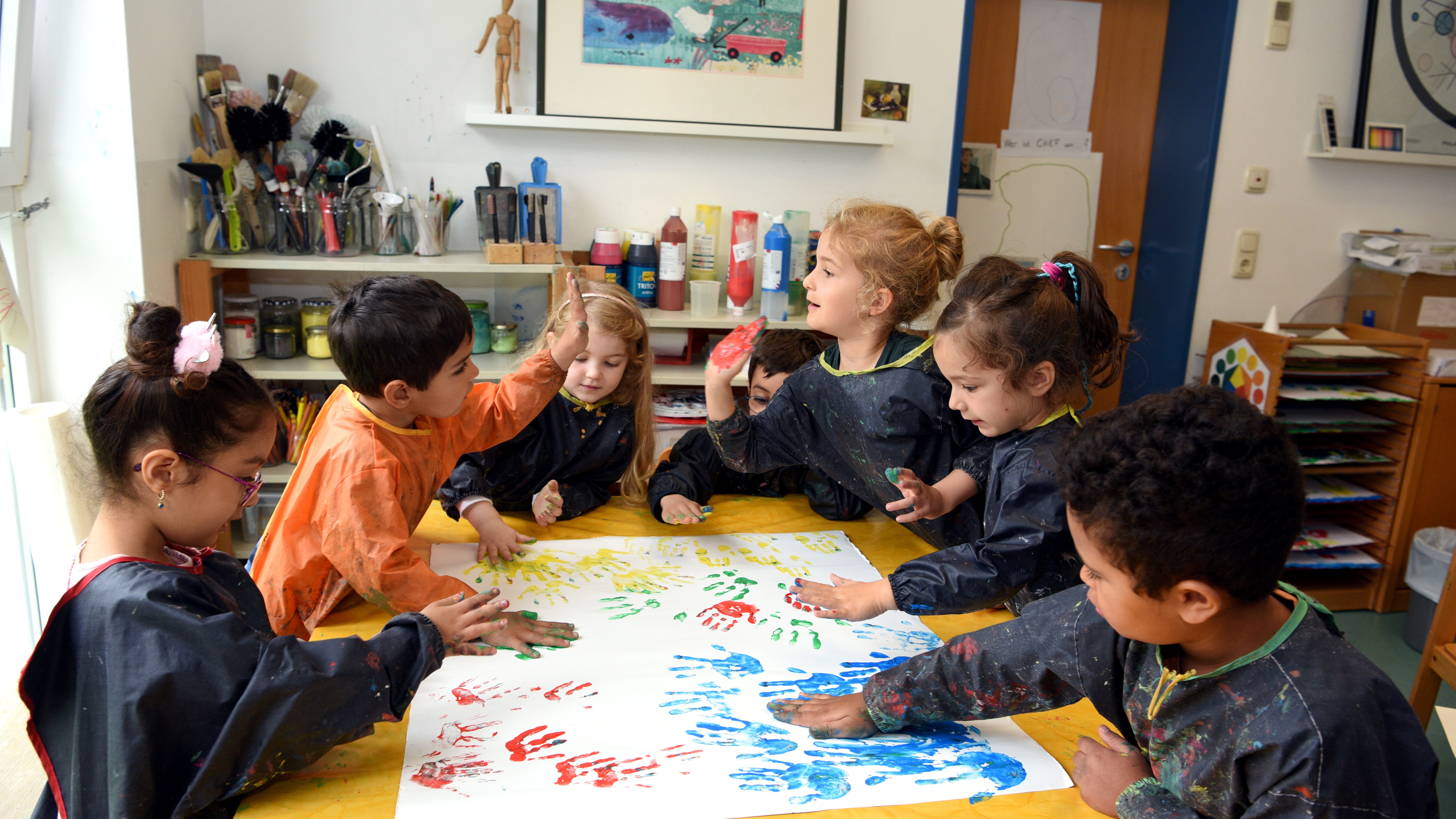 Sechs Kindergartenkinder sitzen um einen Tisch im Kindergarten. Auf dem Tisch liegt ein großes Papier. Darauf machen die Kinder mit Fingerfarben Handabdrücke.