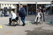 2 Menschen sitzen im Rollstuhl und werden von einer anderen Person durch einen Hütchen-Parcours geschoben