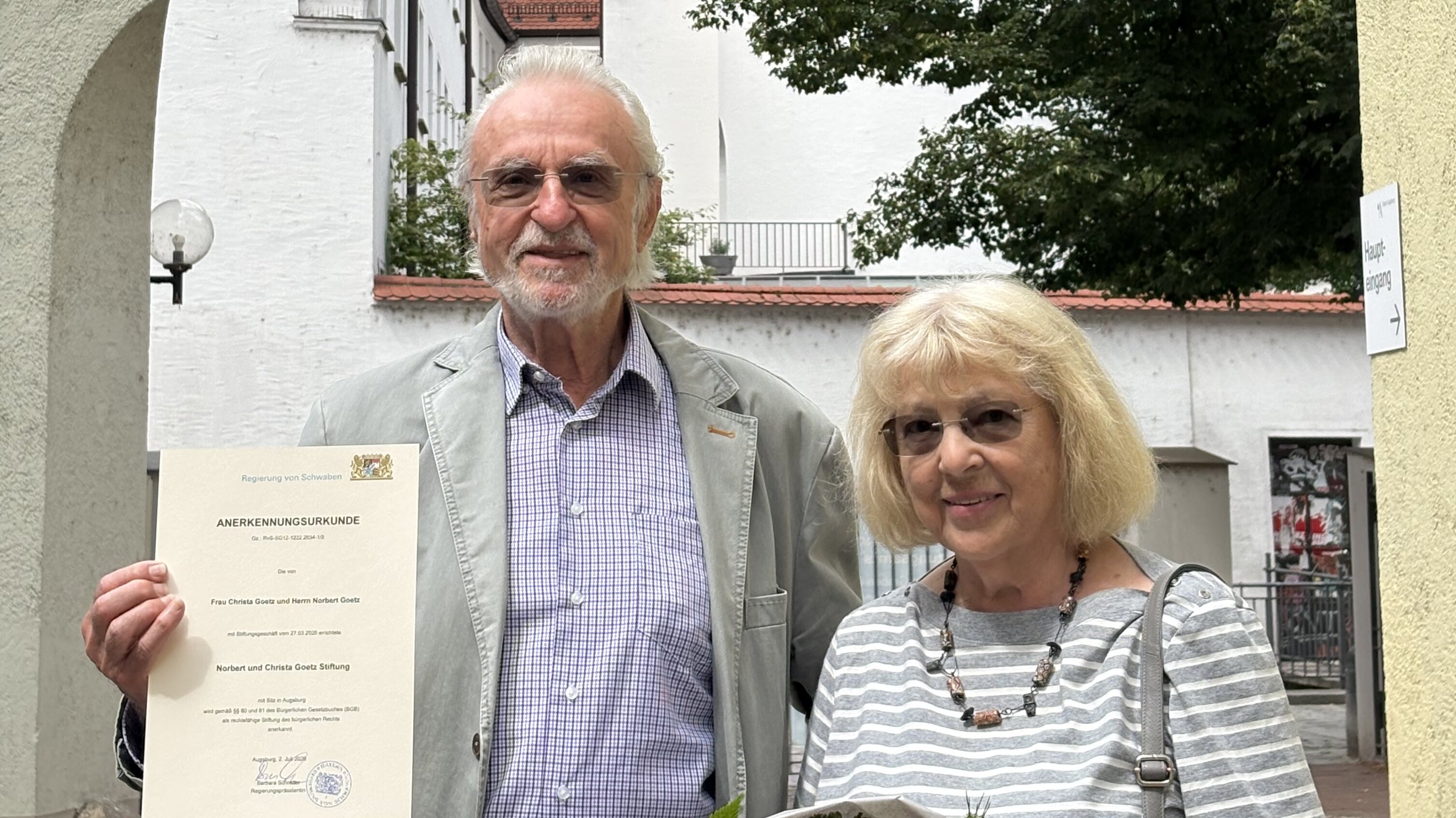 ein älteres Paar vor historischem Gebäude; er hält eine Stiftungsurkunde hoch
