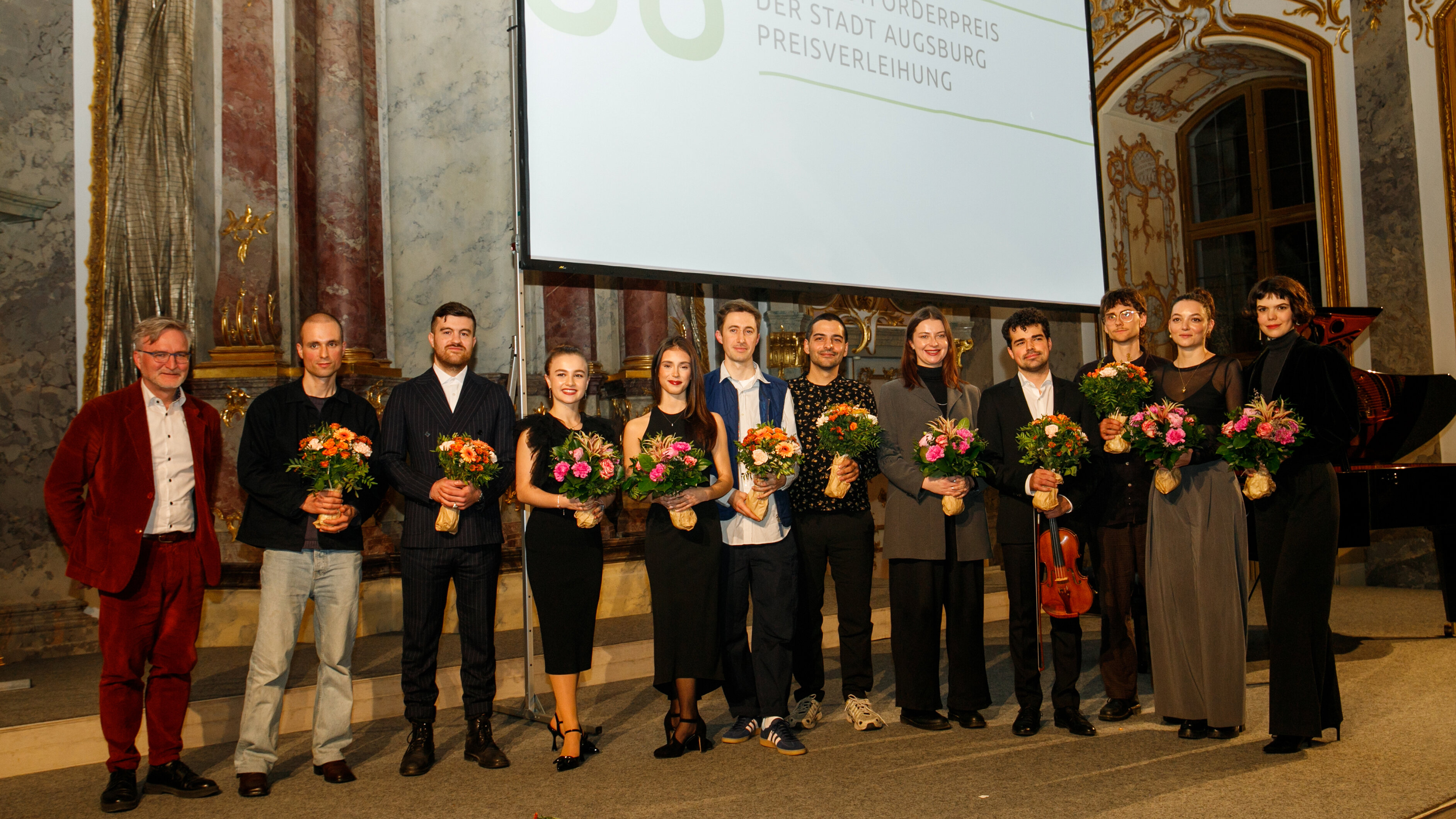 Gruppenbild mit festlich gekleideten Personen beiderlei Geschlechts vor einer gro0en Leinwand; alle halten einen Blumenstraußarauf 