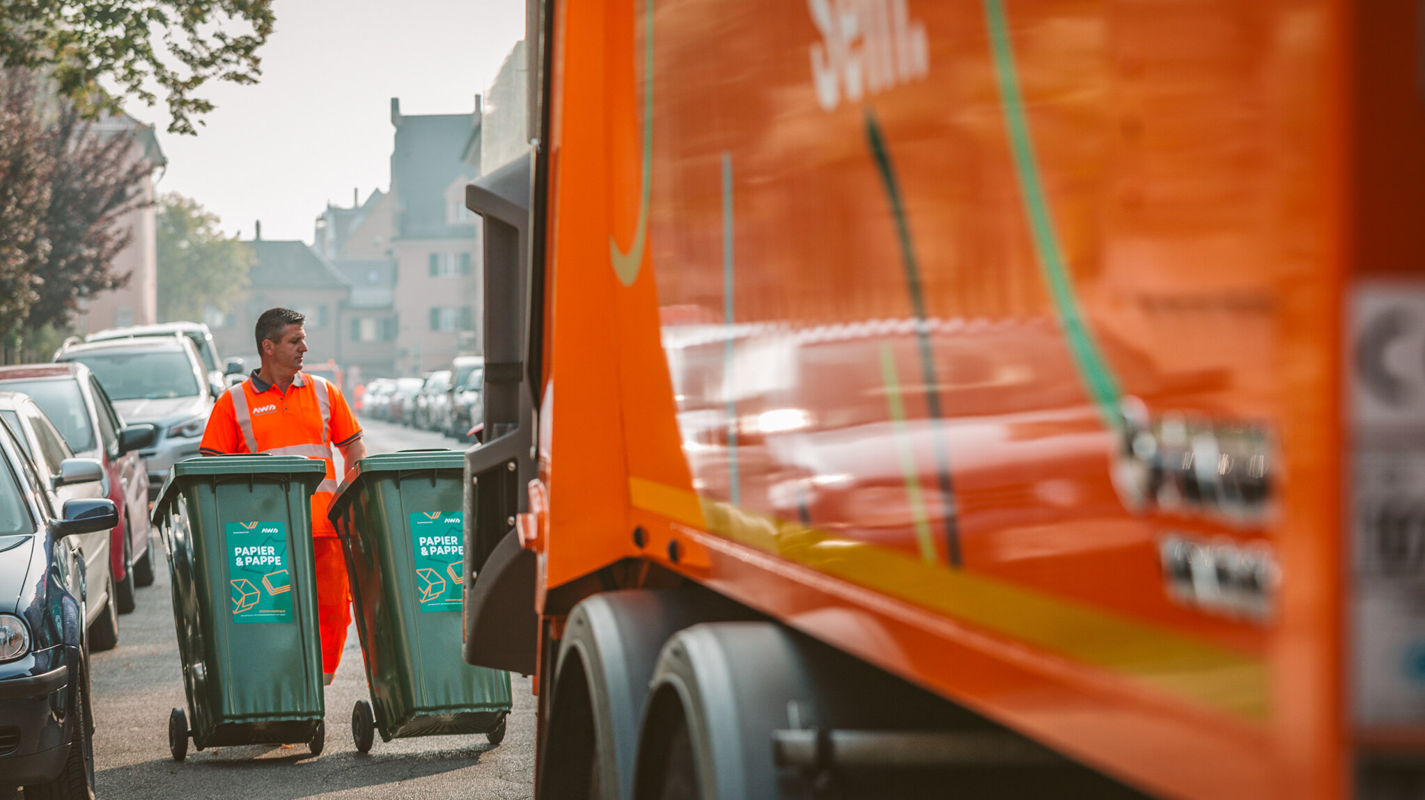 Müllfahrzeug von der Seite; dahinter ein Mann in Arbeitskleidung, der zwei Papiertonnen zum Fahrzeug schiebtebt