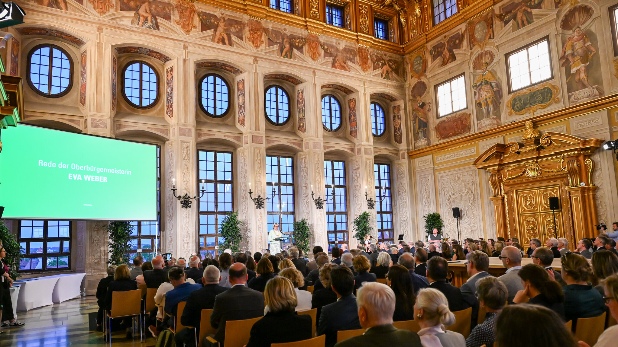 Viele Menschen sitzen in einem großen Saal, die Wände sind mit Gold verziert