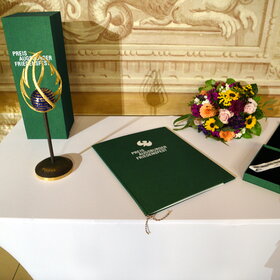 Auf dem Tisch steht links eine Skulptur. In der Mitte liegt eine Urkunde und dahinter steht ein Blumenstrauß. Rechts ist eine Medaille zu sehen.