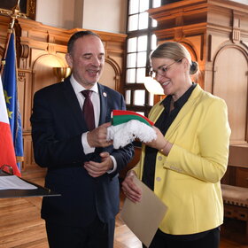 Eine Frau übergibt einem Mann einen Schal. Im Hintergrund ist die französische Flagge zu sehen.