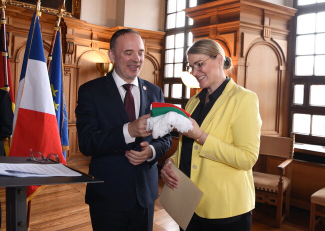 Eine Frau übergibt einem Mann einen Schal. Im Hintergrund ist die französische Flagge zu sehen.