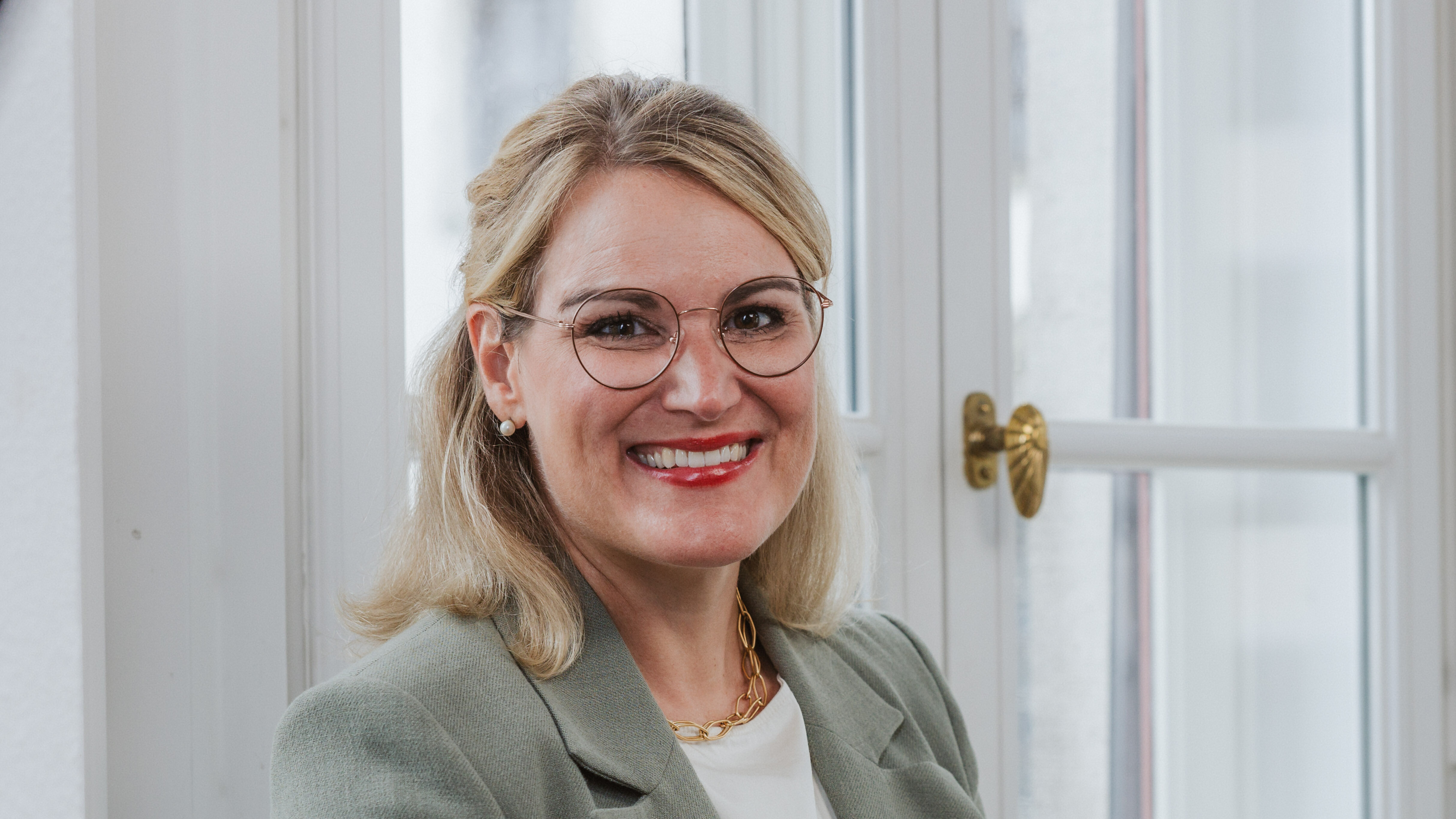 Frau mit schulterlangem Haar, Brille und Sakko lehnt an Fensterkreuz