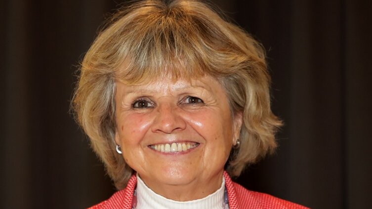 Portraitbild einer lächelnden Frau mit halblangem Haar, die einen Rollkragenpullover, eine Strickjacke und eine längere Halskette trägt