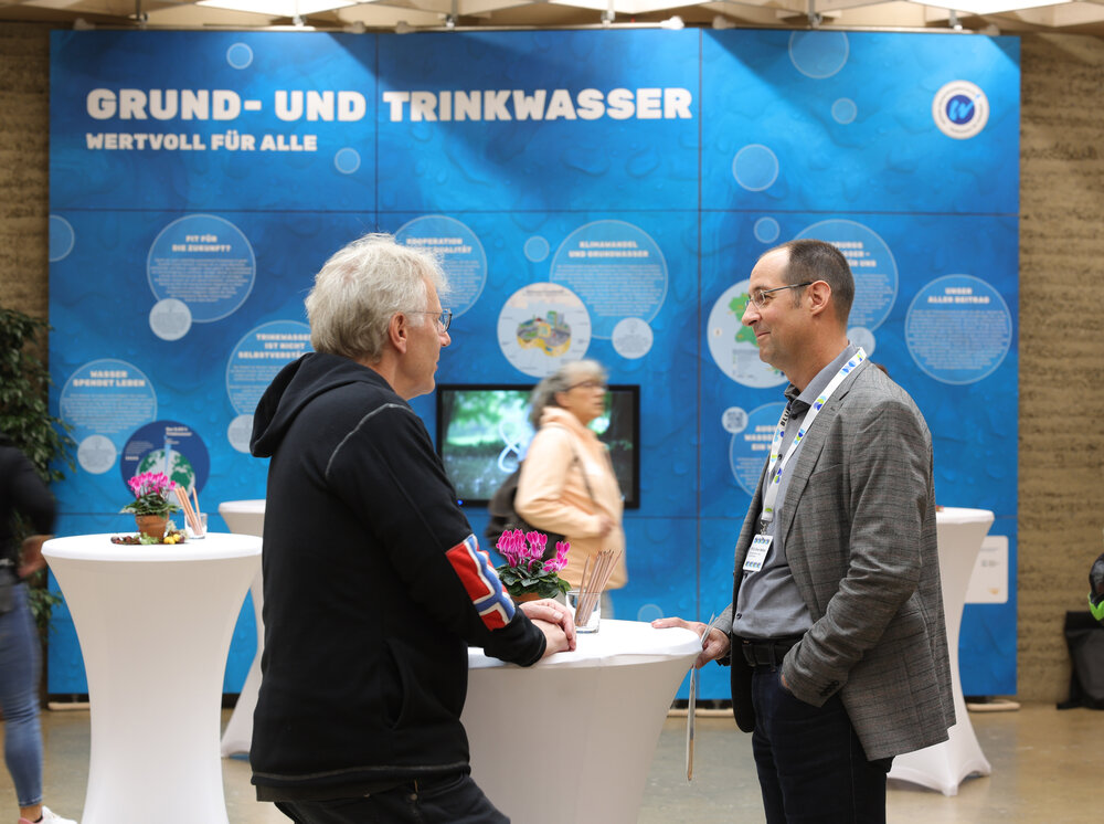 Zwei Männer unterhalten sich an einem Stehtisch. Im Hintergrund ist eine große Werbewand mit der Aufschrift Grund- und Trinkwasser zu sehen.