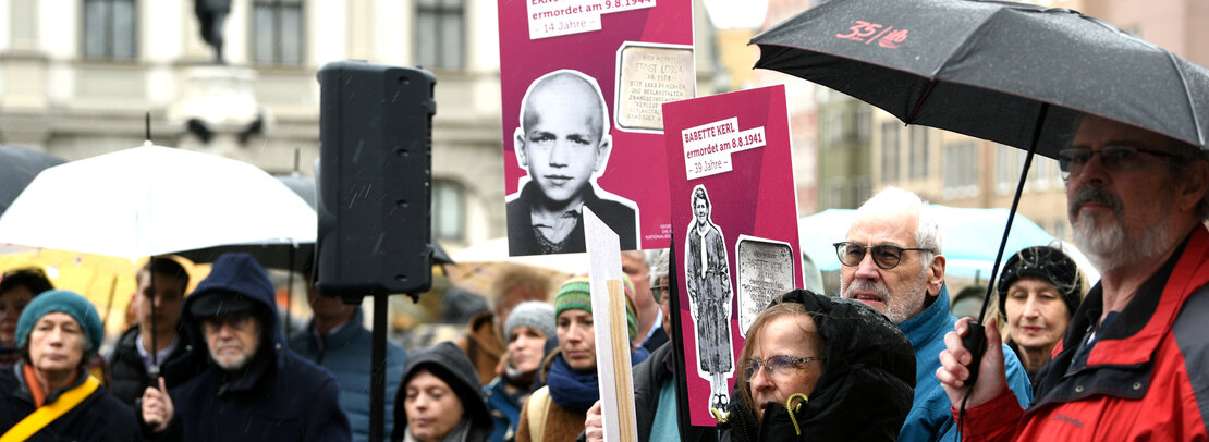 Menschen mit Kapuzen und Regenschirmen halten Plakate hoch mit Fotos eines Jungen bzw. einer Frau, darauf deren Name und der Zusatz "ermordet am ... xx Jahre"