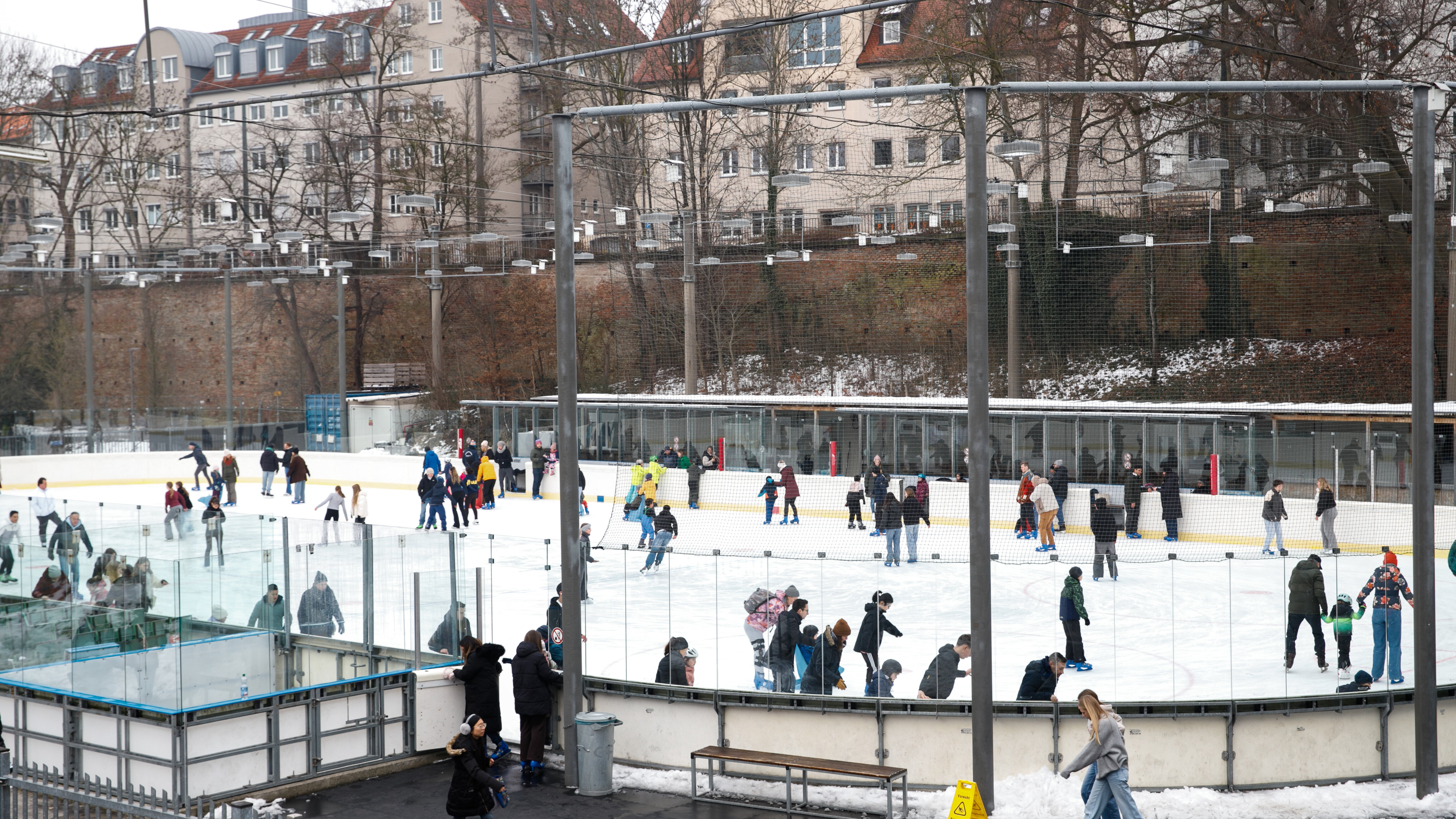 Eine Eisbahn ohne Dach. Es sind viele Leute beim Schlittschuhlaufen. Im Hintergrund ist eine Stadtmauer zu sehen