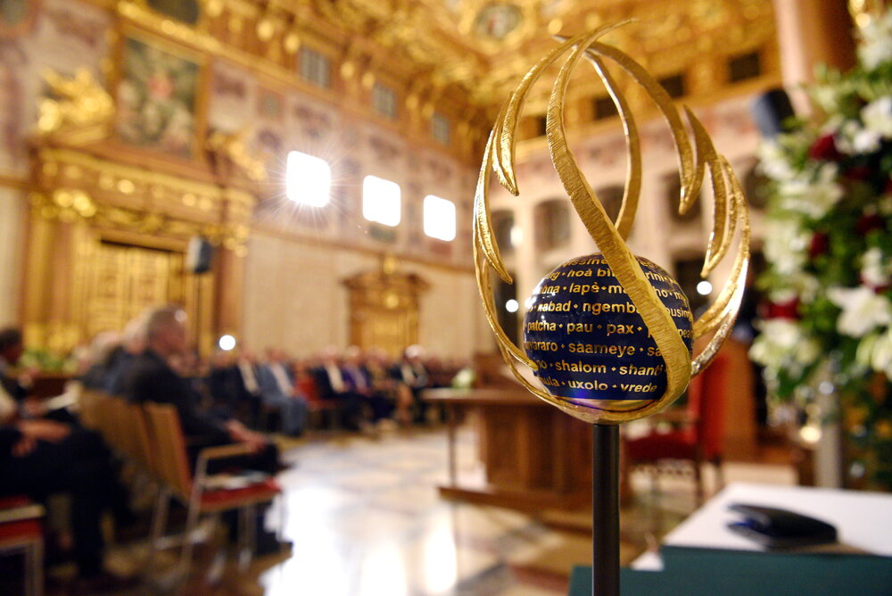 Im Vordergrund steht eine Skulptur. Im Hintergrund ist ein Saal mit sitzenden Gästen zu sehen.