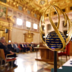 Im Vordergrund steht eine Skulptur. Im Hintergrund ist ein Saal mit sitzenden Gästen zu sehen.