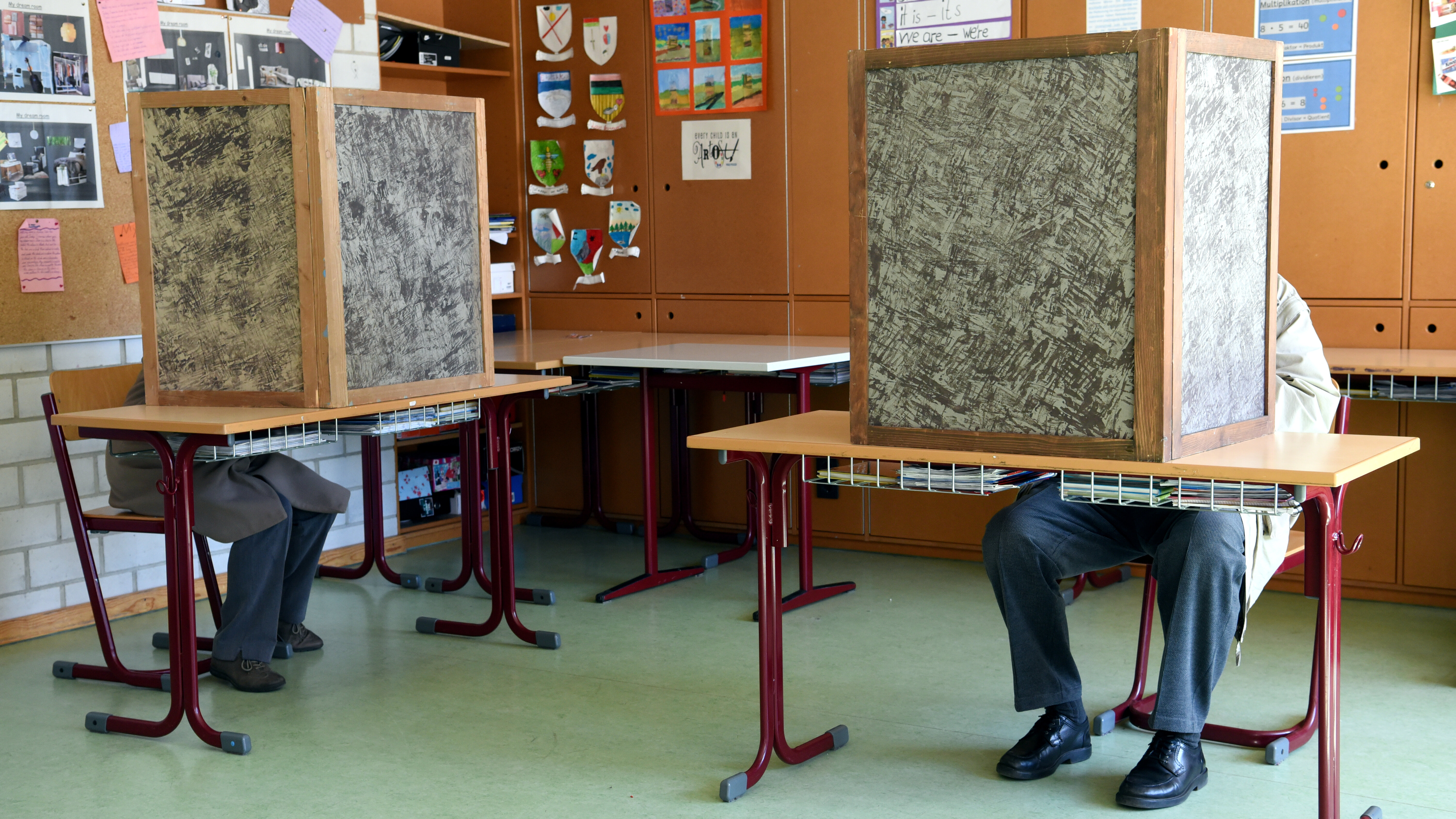 Das Bild zeigt zwei Personen beim Wählen in einem Wahllokal. Sie sitzen an Tischen. Um sie herum ist eine Wahlkabine.
