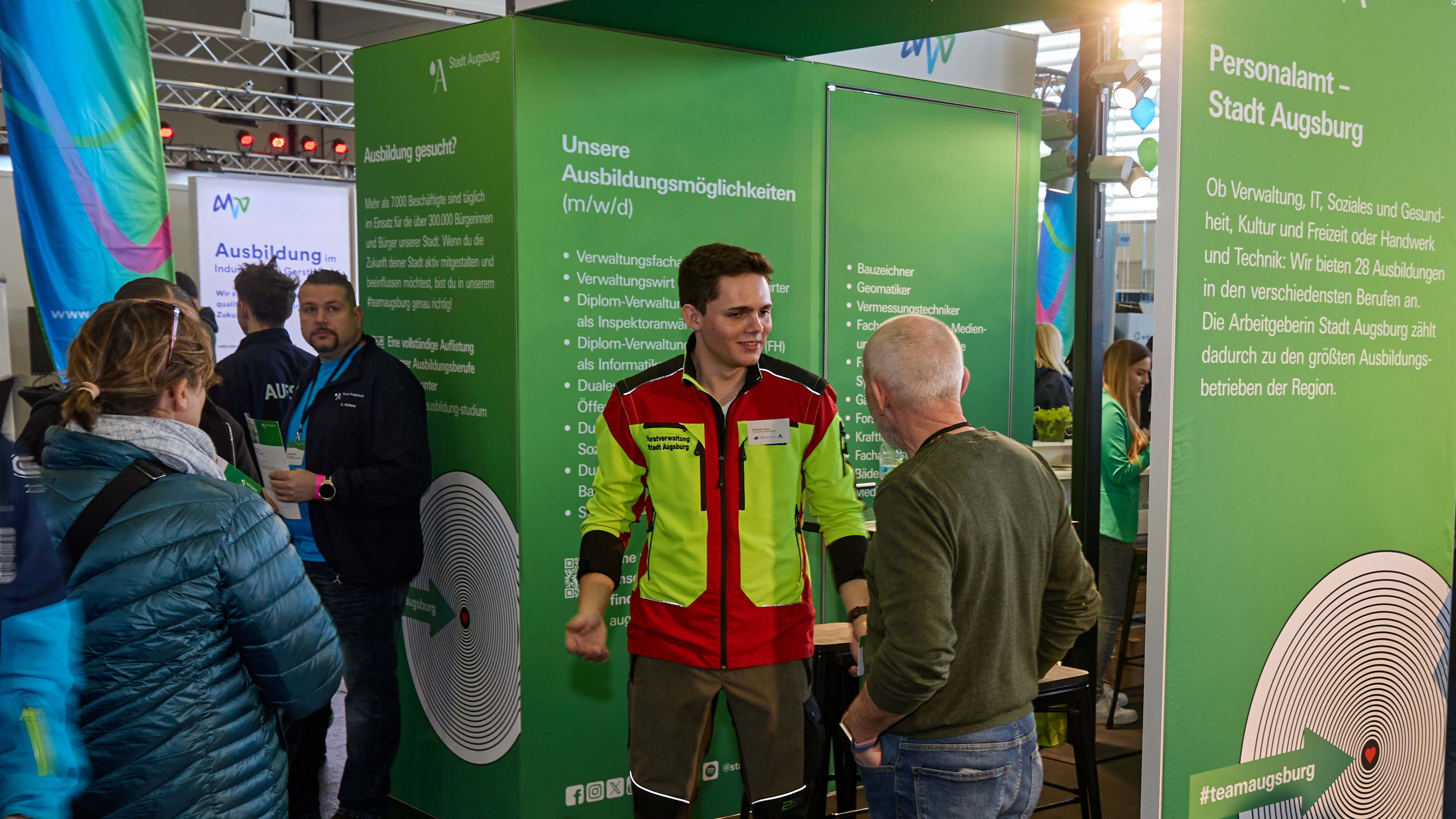 Ein Stand auf einer Messe. Ein junger Mann in Förster-Kleidung erzählt einem anderen Mann etwas