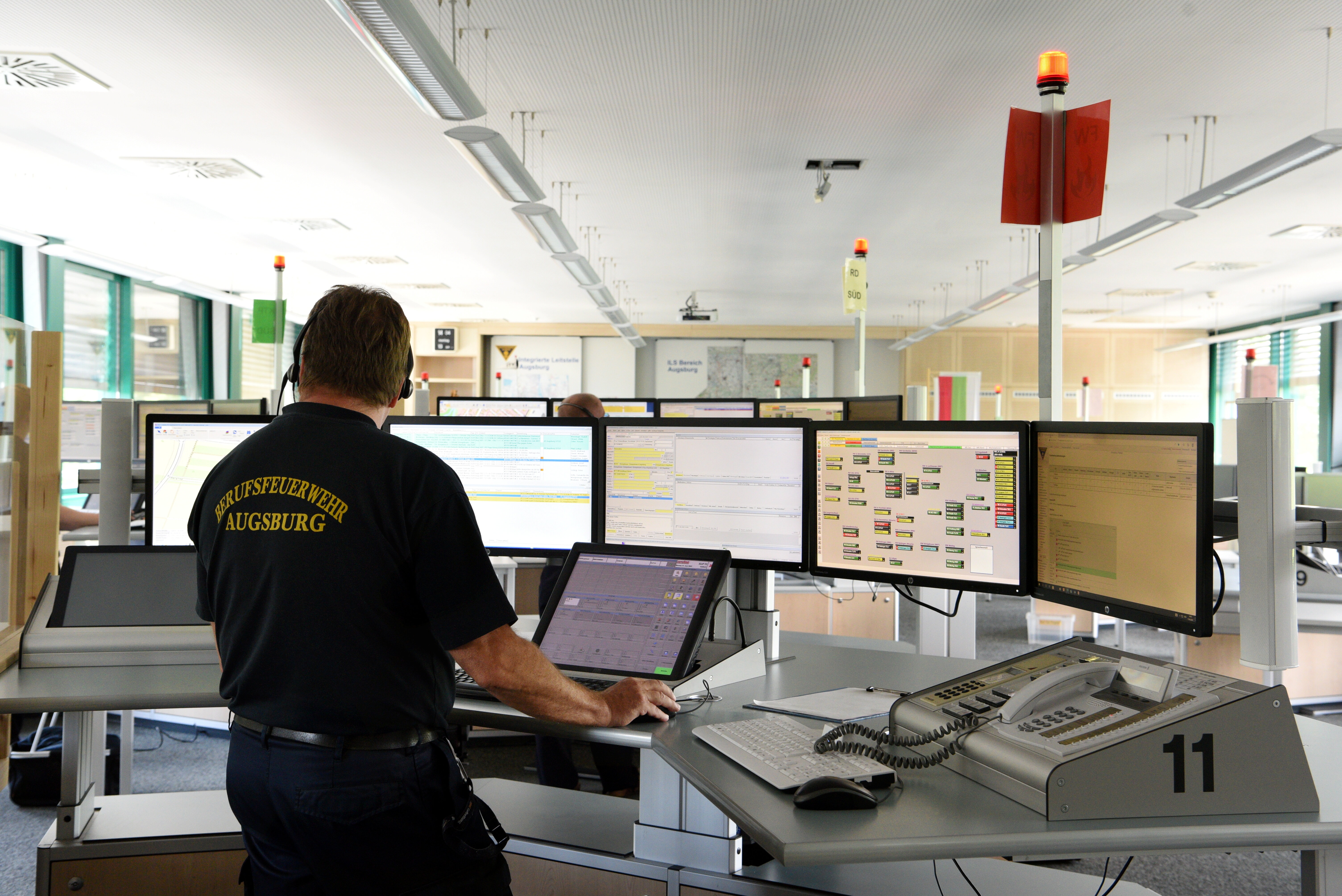 Ein Mann mit der Aufschrift "Berufsfeuerwehr Augsburg" auf seinem Hemd steht vor sechs Bildschirmen.