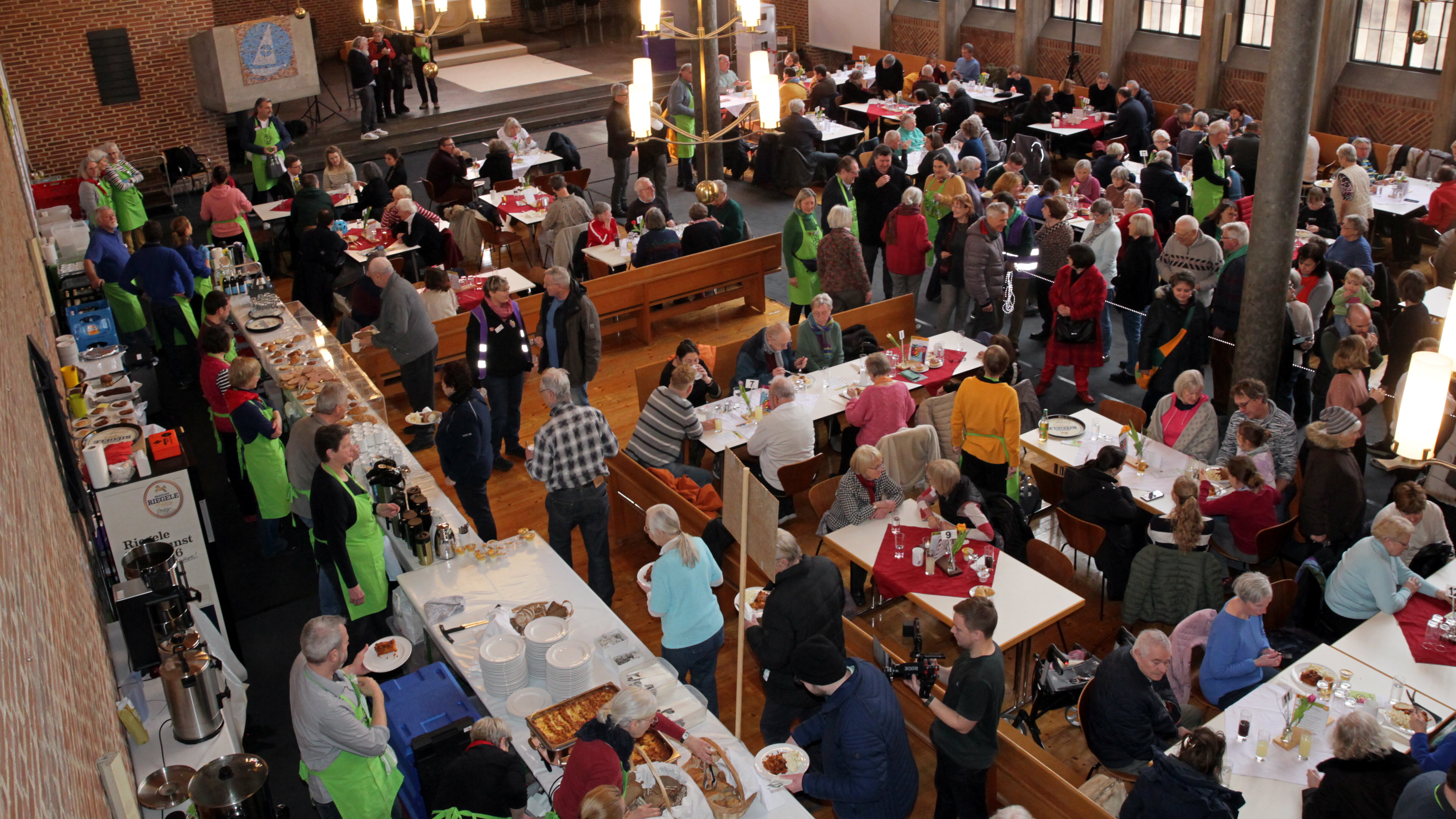 Das Foto zeigt den Innenraum einer Kirche. An der Seite sind Tische aneinander gereiht, an denen Essen von Menschen, die grüne Schürzen trage, ausgegeben wird. An vielen anderen Tische, sitzen Menschen und essen.