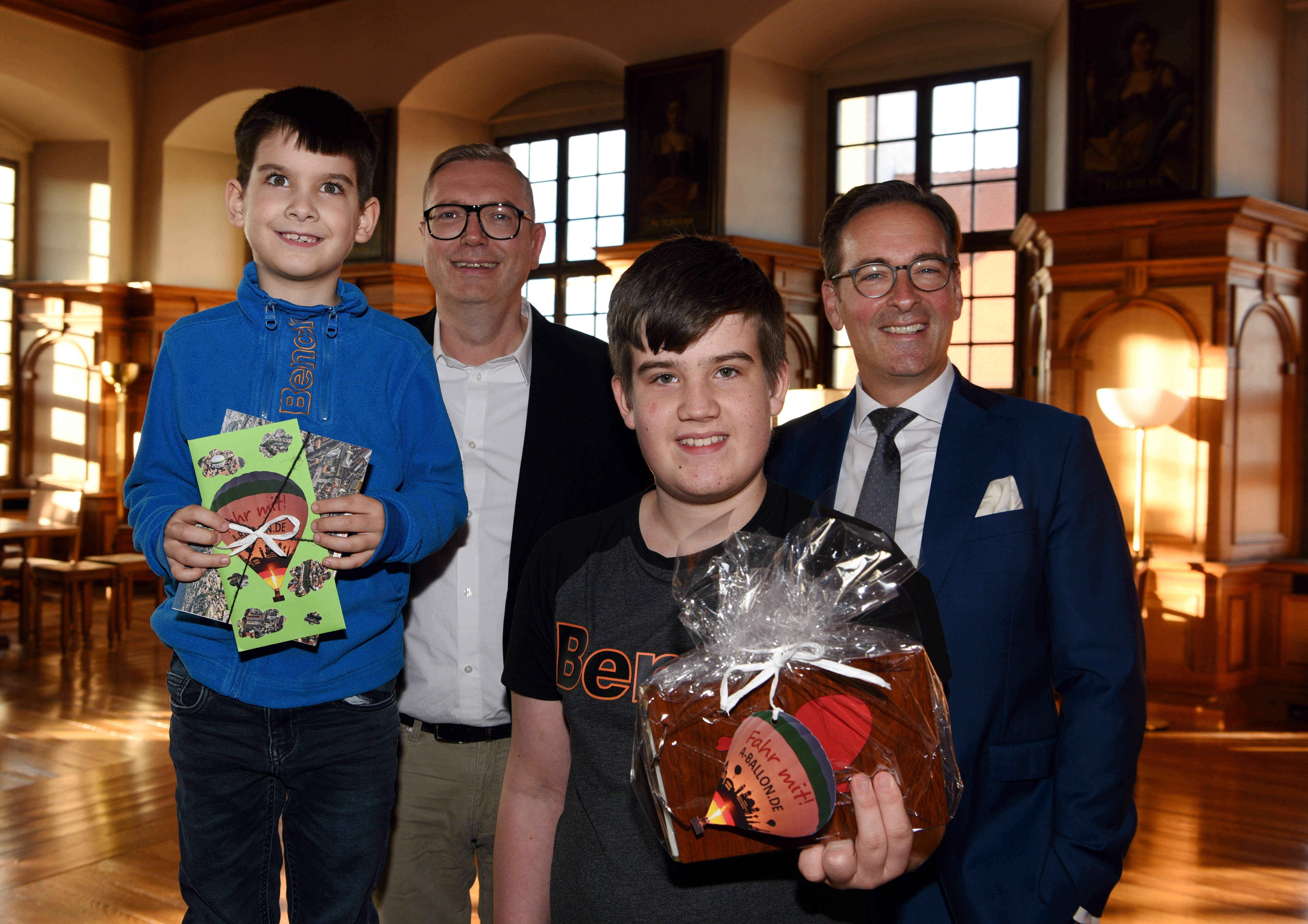 Zwei lachende Kinder und zwei erwachsene Männer in einem mit Holz dekorierten Raum des Rathauses. Die Kinder haben beim Turamichele einen Preis gewonnen. Die beiden Männer übergeben den Kindern den Preis.