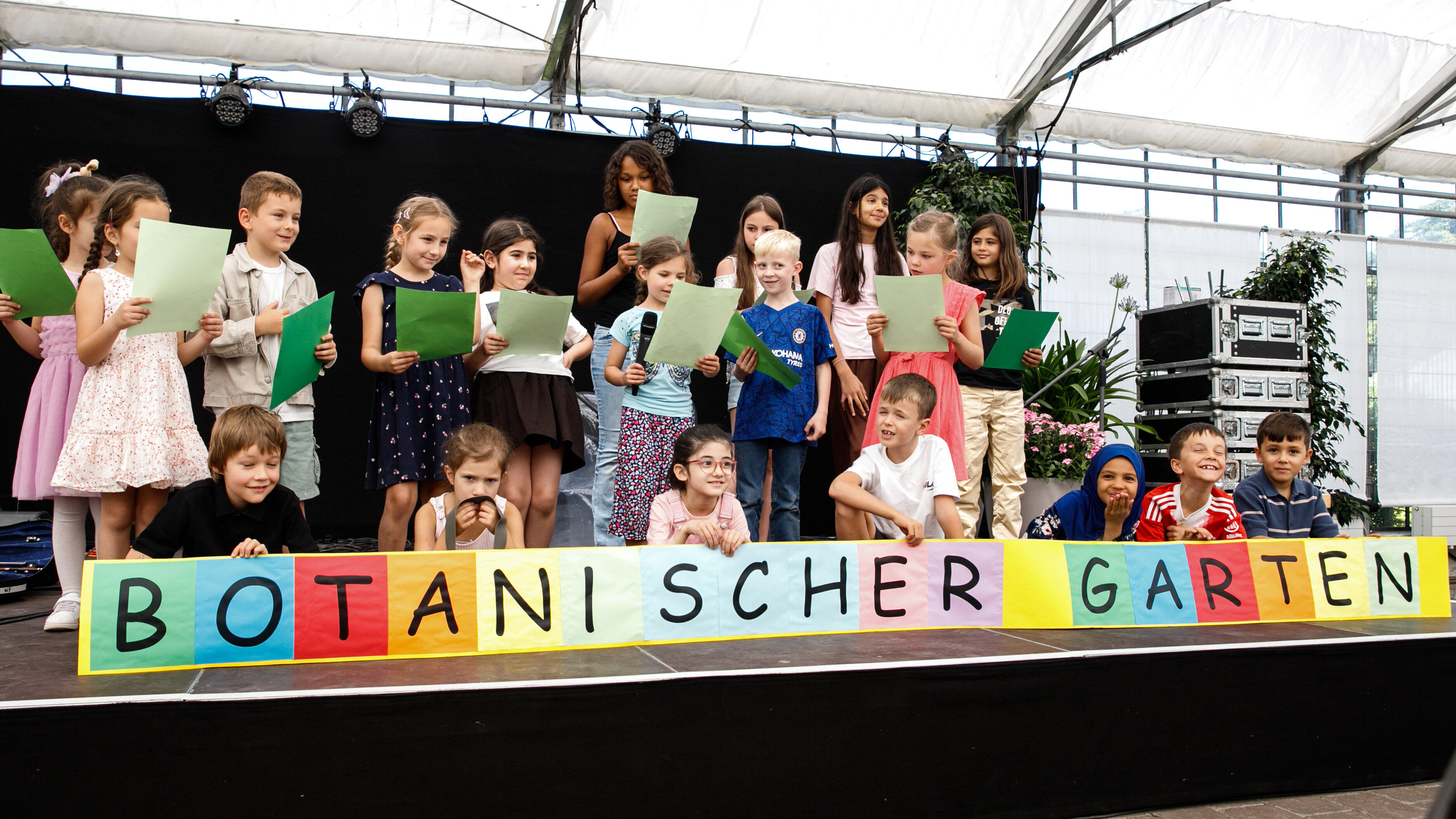Viele Kinder auf einer Bühne. Darunter steht in bunten Buchstaben "Botanischer Garten"
