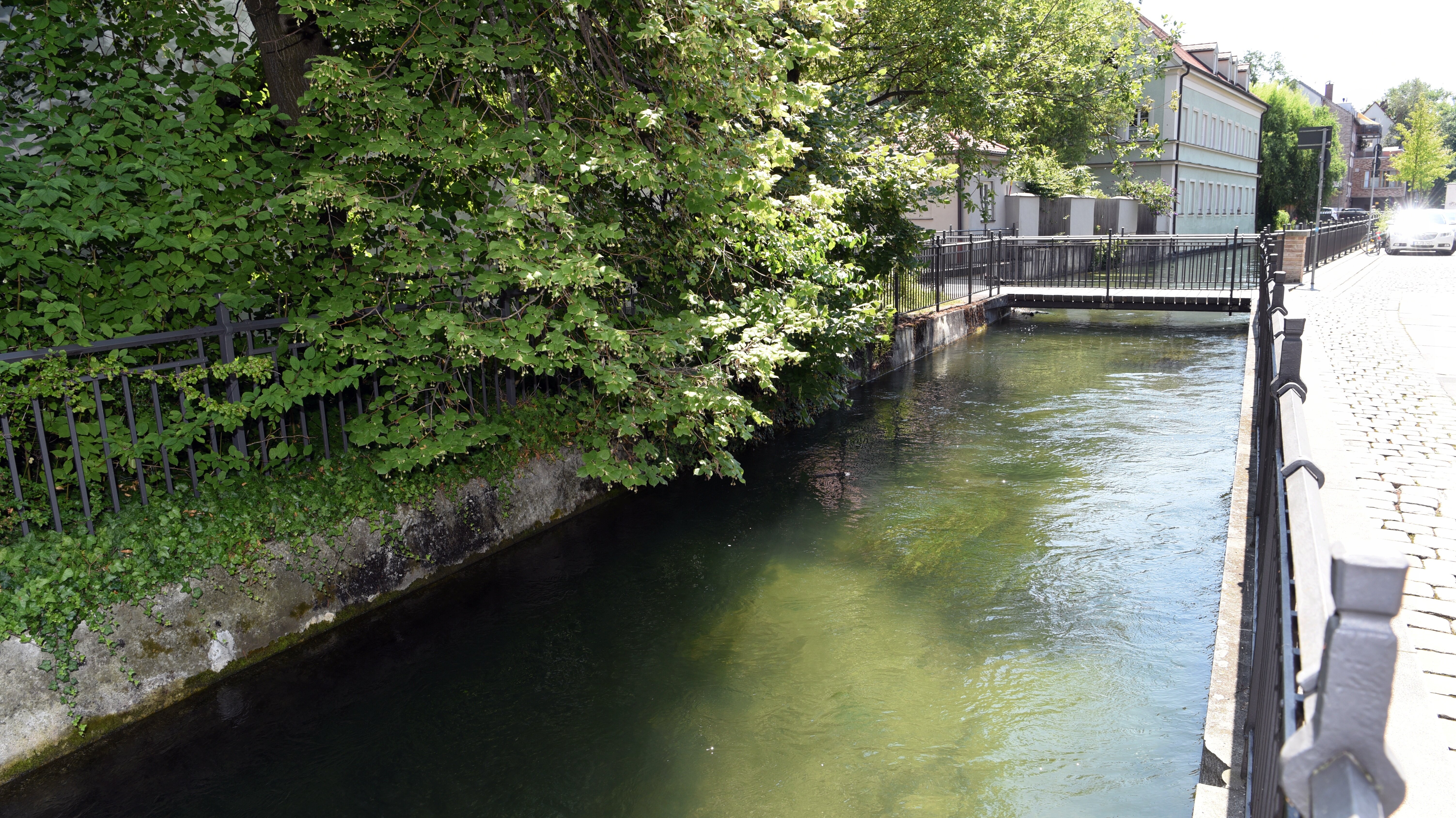 ein Kanal, der links von Bäumen gesäumt ist. Rechts befindet sich ein Geländer und eine Straße mit Kopfsteinpflaster