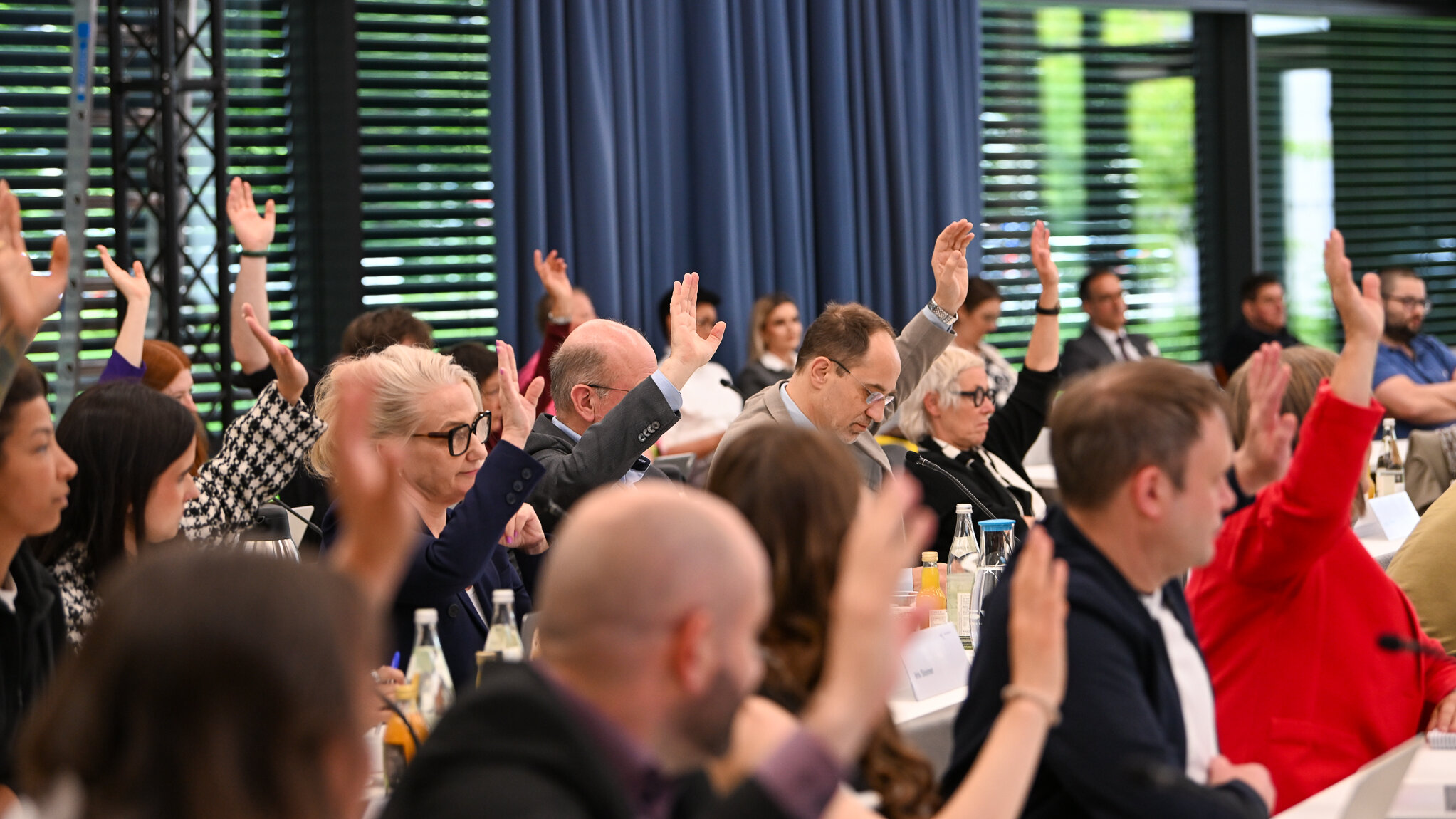 Viele Menschen sitzen an Tischen in einem Saal. Viele heben die Hand zu einer Abstimmung