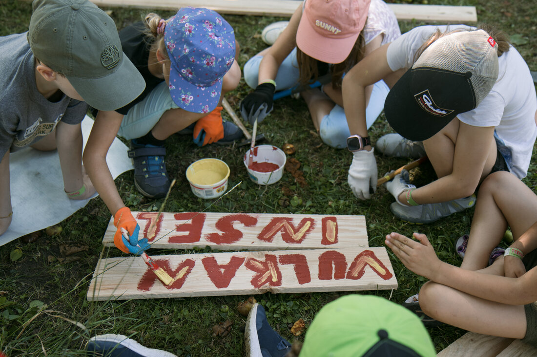 mehrere Kinder sitzen auf dem Boden und bemalen Holzschilder mit rotem und gelben Schriftzug: "Vulkaninsel"