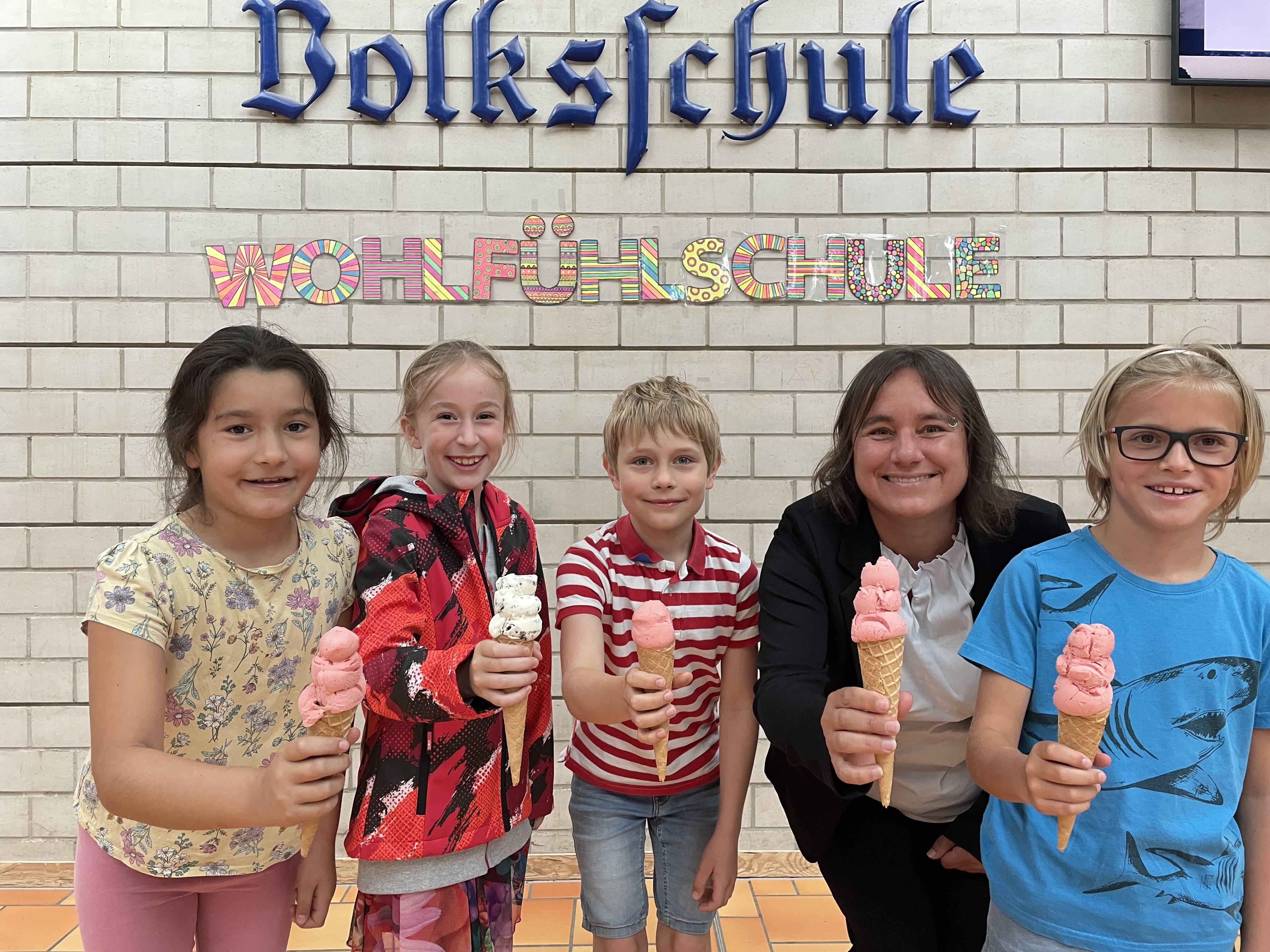 Eine Frau und vier Kinder haben Eiswaffeln in der Hand. Im Hintergrund an einer Wand steht "Volksschule" und "Wohlfühlschule"