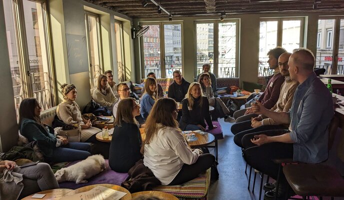 in einem Café sitzen ca 20 Menschen an kleinen Tischen vor 3 Männern auf Barhockern und hören zu, was diese erzählen.