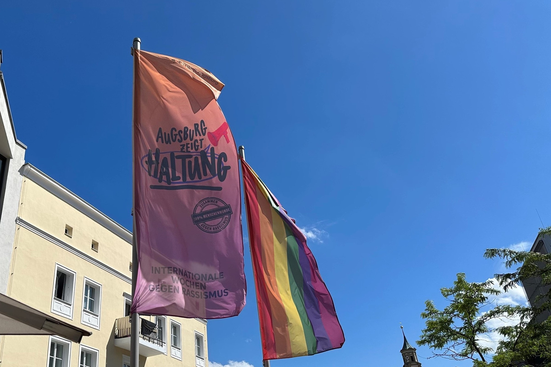 Zwei Flaggen im Wind vor blauen Himmel. Links die der Wochen gegen Rassismus, rechts eine Regenbogenflagge.