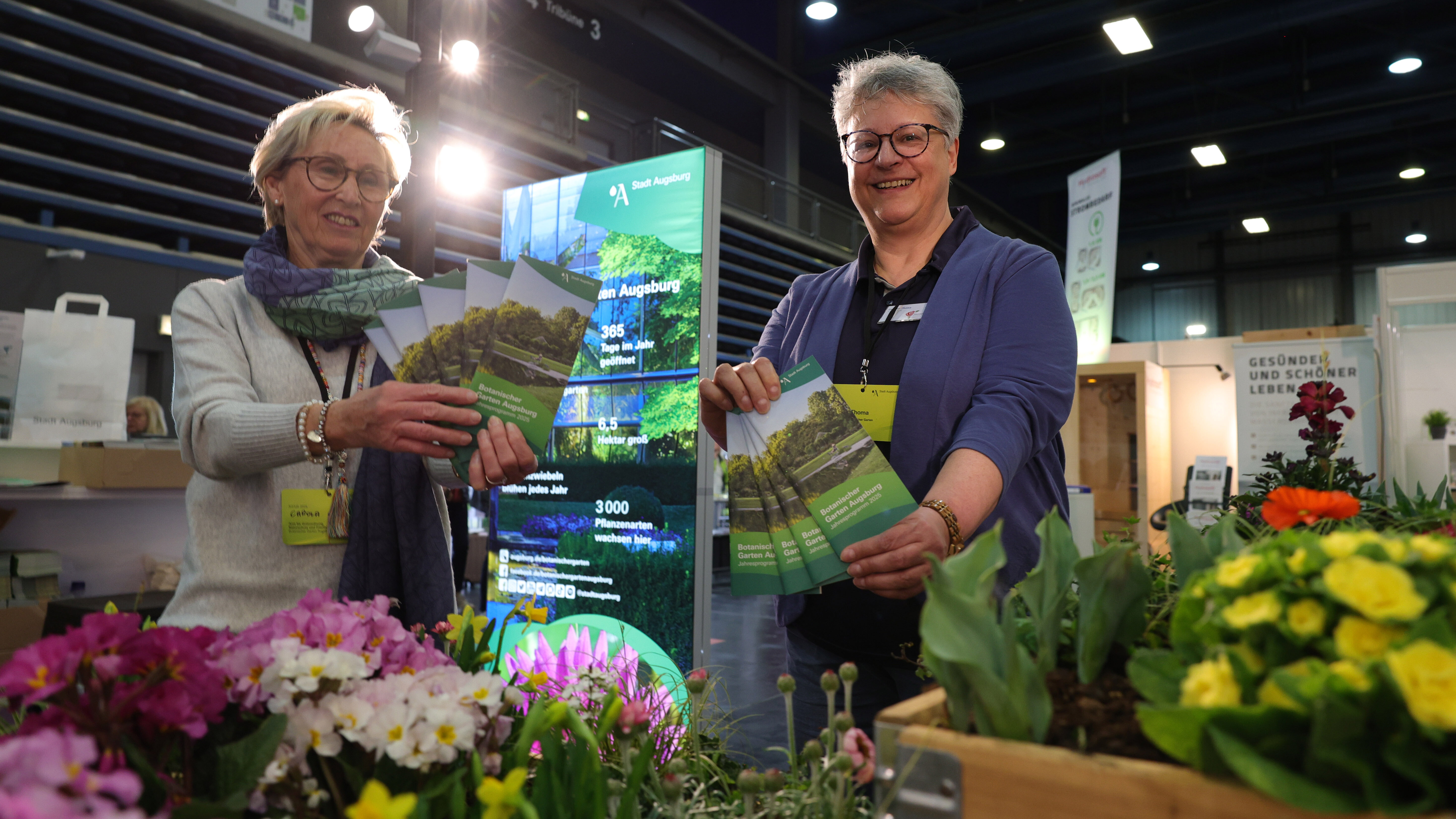 Zwei Frauen halten Flyer vom Botanischen Garten in die Kamera. Im Vordergrund sind Blumen zu sehen, im Hintergrund ein Plakat auf dem Stadt Augsburg steht. 