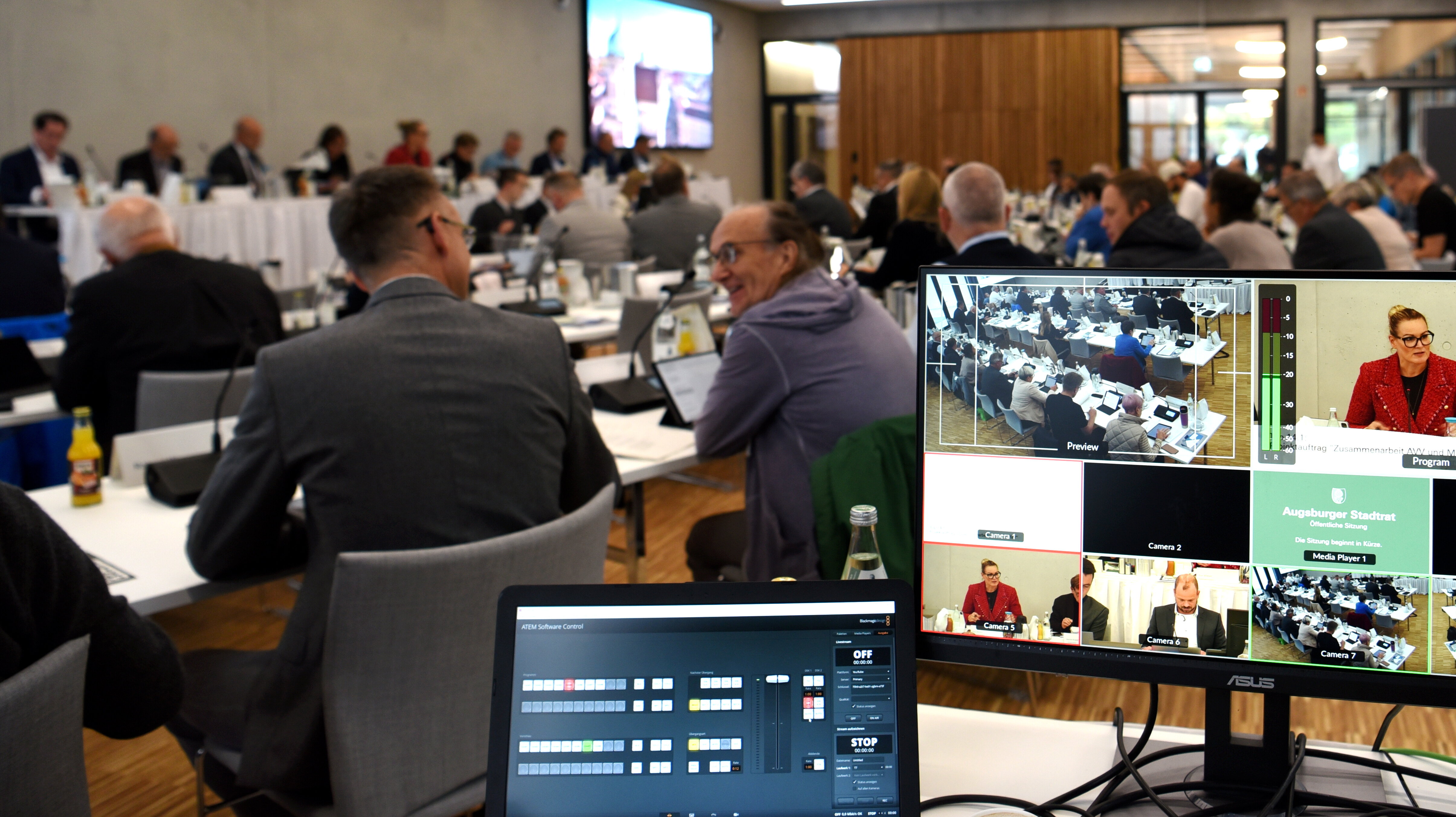 Viele Menschen sitzen an langen Tischen und diskutieren. Im Vordergrund ein Bildschirm, auf dem die Sitzung übertragen wird.