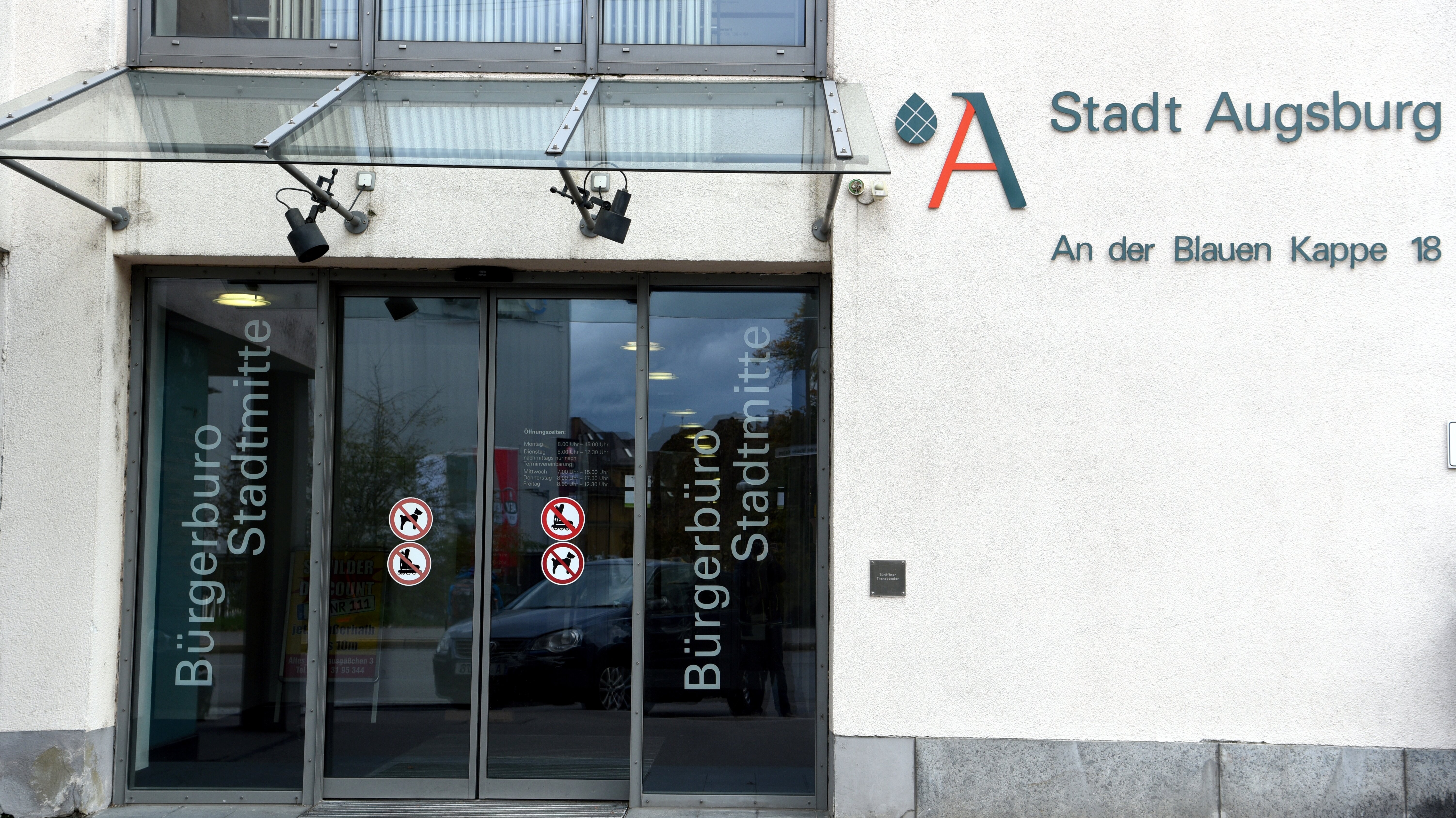 Eingangsbereich des Bürgerbüros Stadtmitte mit gläsernen Schiebetüren, auf denen vier Verbotsschilder angebracht sind, darüber ein gläsernes Vordach, rechts an der Mauer das Logo und der Schriftzug der Stadt Augsburg