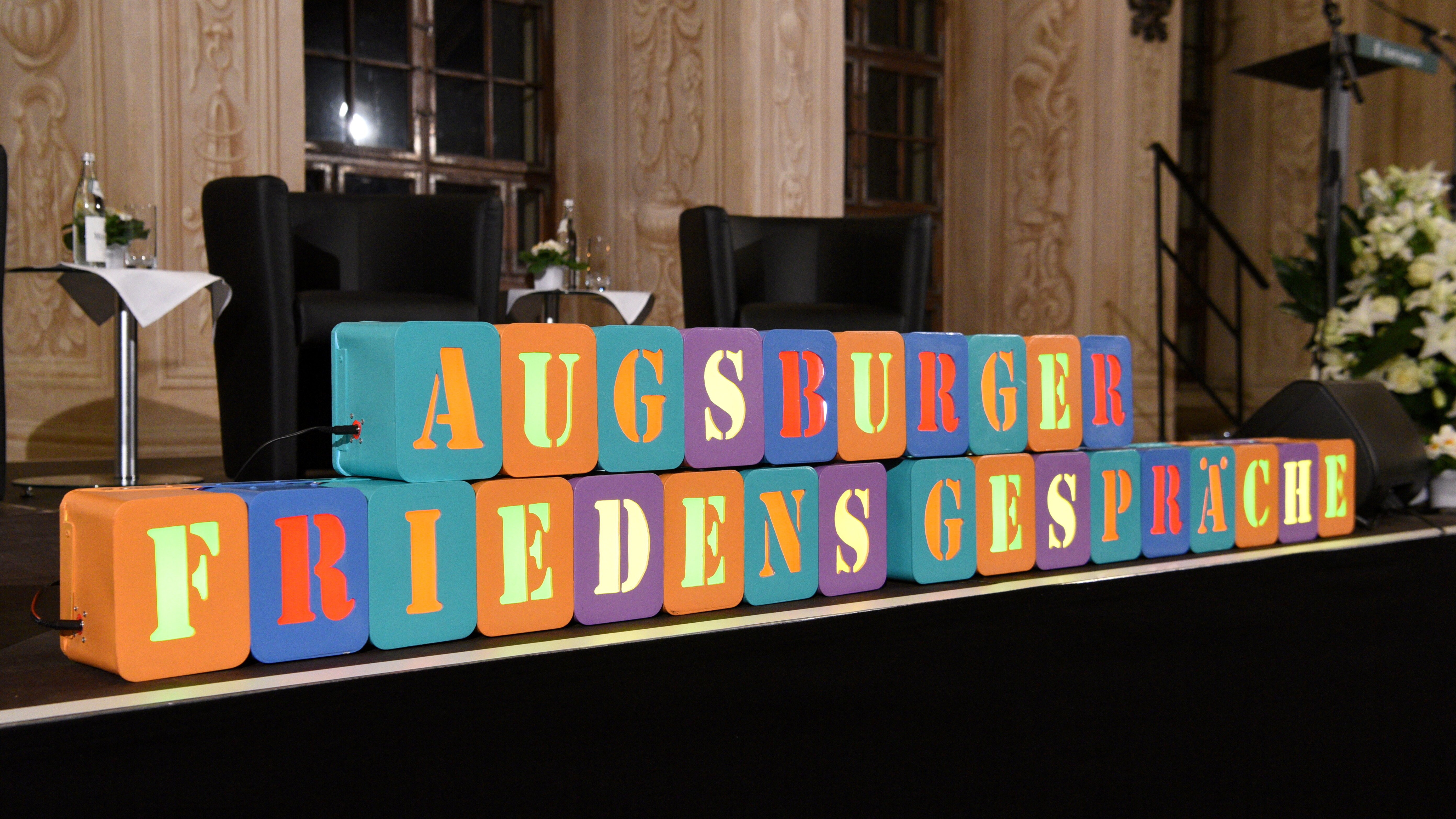 Das Foto zeigt eine Bühne im Goldenen Saal des Augsburger Rathauses. Darauf stehen zwei Stühle. Im Vordergrund stehen bunte Würfel mit Buschtaben darauf. Diese schreiben "Augsburger Friedensgespräche"