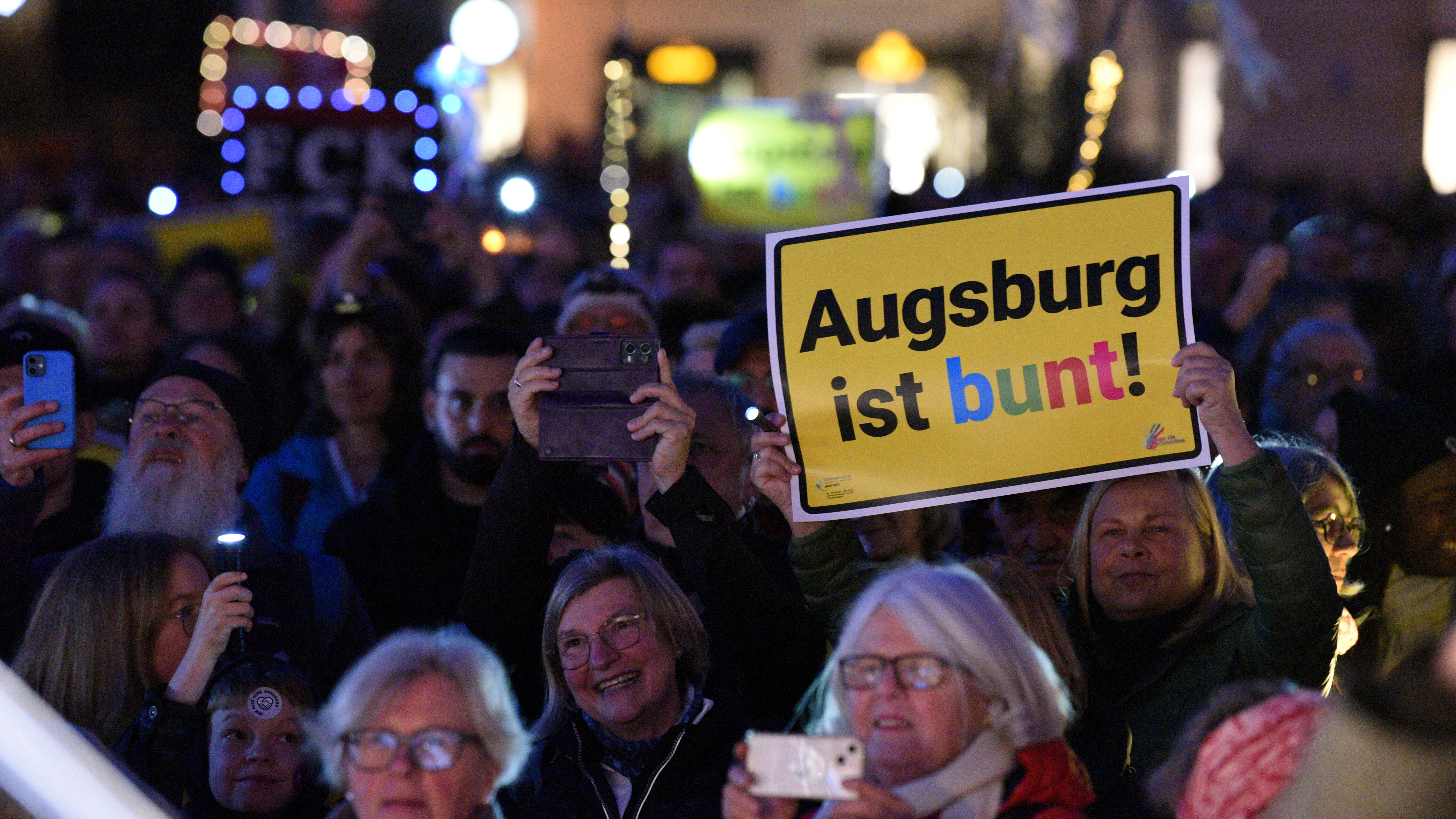 nächtliche Demonstration; viele Menschen nutzen ihre Handy-Taschenlampe; im Vordergrund ein "Ortsschild" mit der Aufschrift "Augsburg ist bunt"