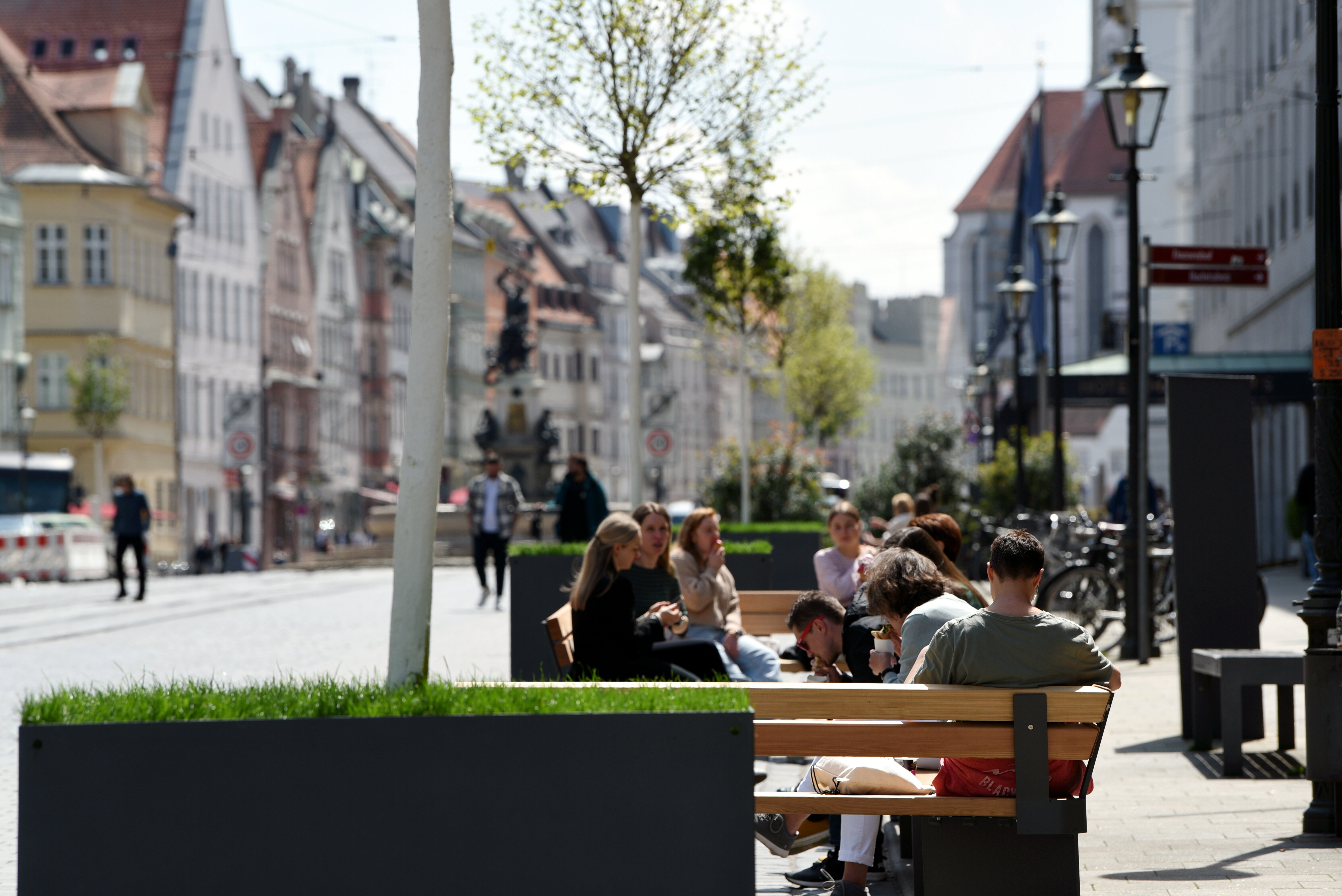 Jüngere Menschen sitzen auf Bänken entlang einer Straße; im Hintergrund Fußgänger