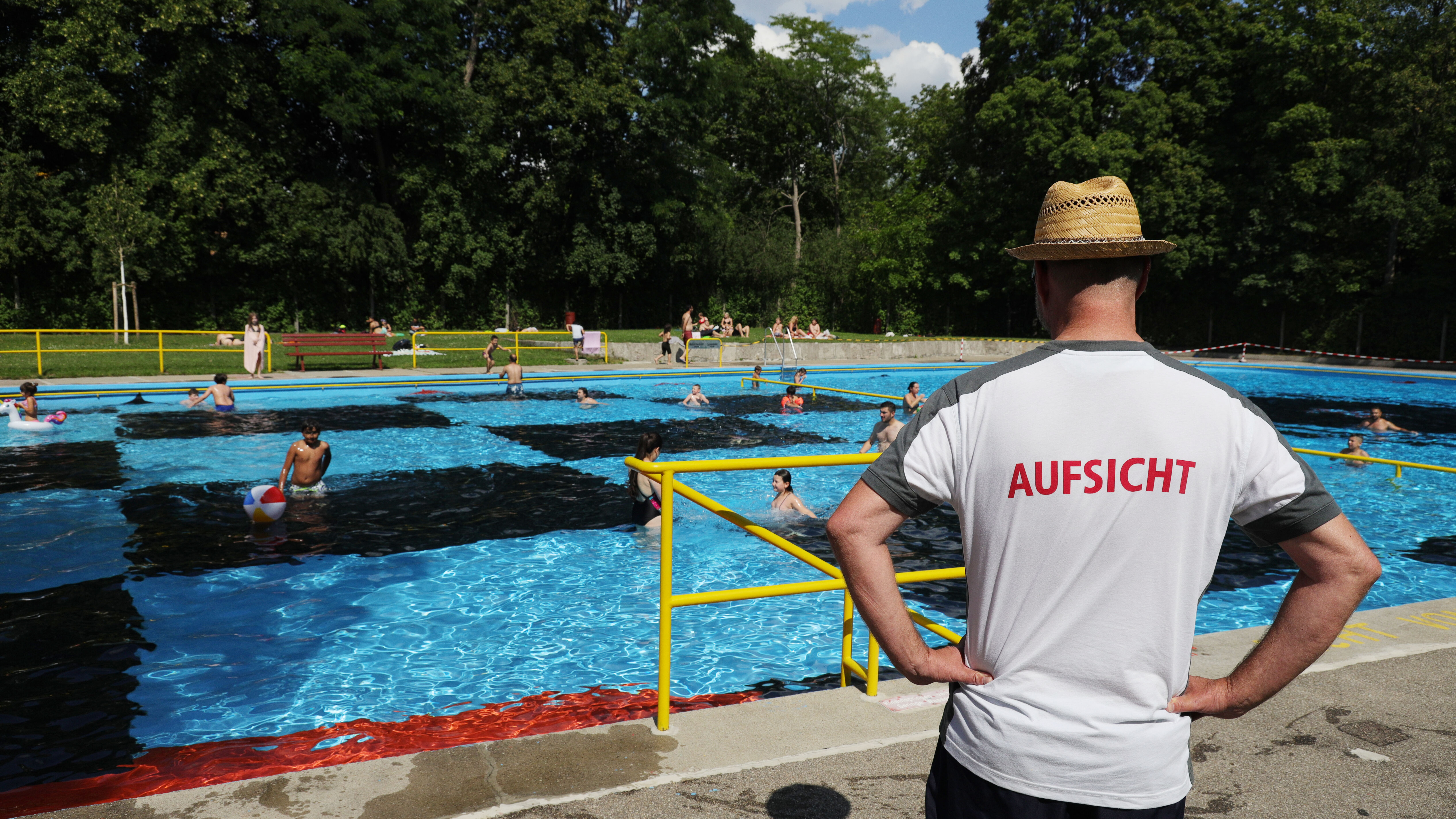 Das Foto zeigt ein Schwimmbecken im Freibad in denen Kinder spielen und Schwimmen. Im Vordergrund des Fotos steht ein Mann, auf dessen T-Shirt Aufsicht steht