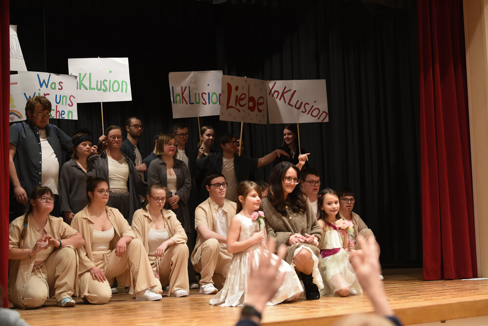 überwiegend junge Menschen knien bzw. stehen auf der Bühne; sie sind hell gekleidet und halten Pappschilder hoch, auf denen "Inklusion" oder "Liebe" steht