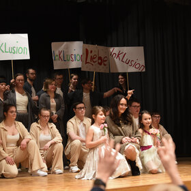 überwiegend junge Menschen knien bzw. stehen auf der Bühne; sie sind hell gekleidet und halten Pappschilder hoch, auf denen "Inklusion" oder "Liebe" steht
