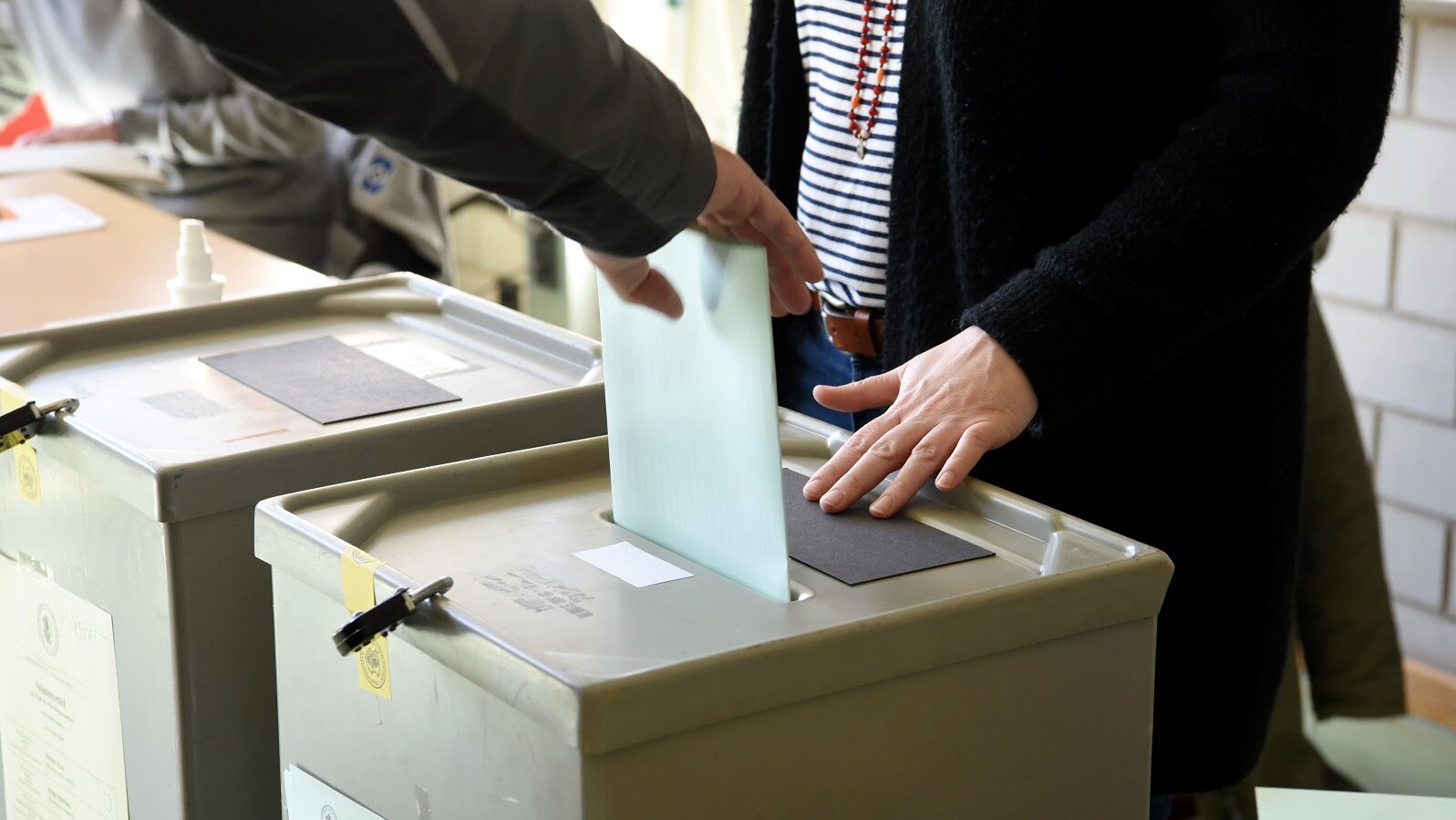 Wahlurne. Eine Person wirft Wahlzettel ein. Eine Person hat ihre Hände auf der Wahlurne.