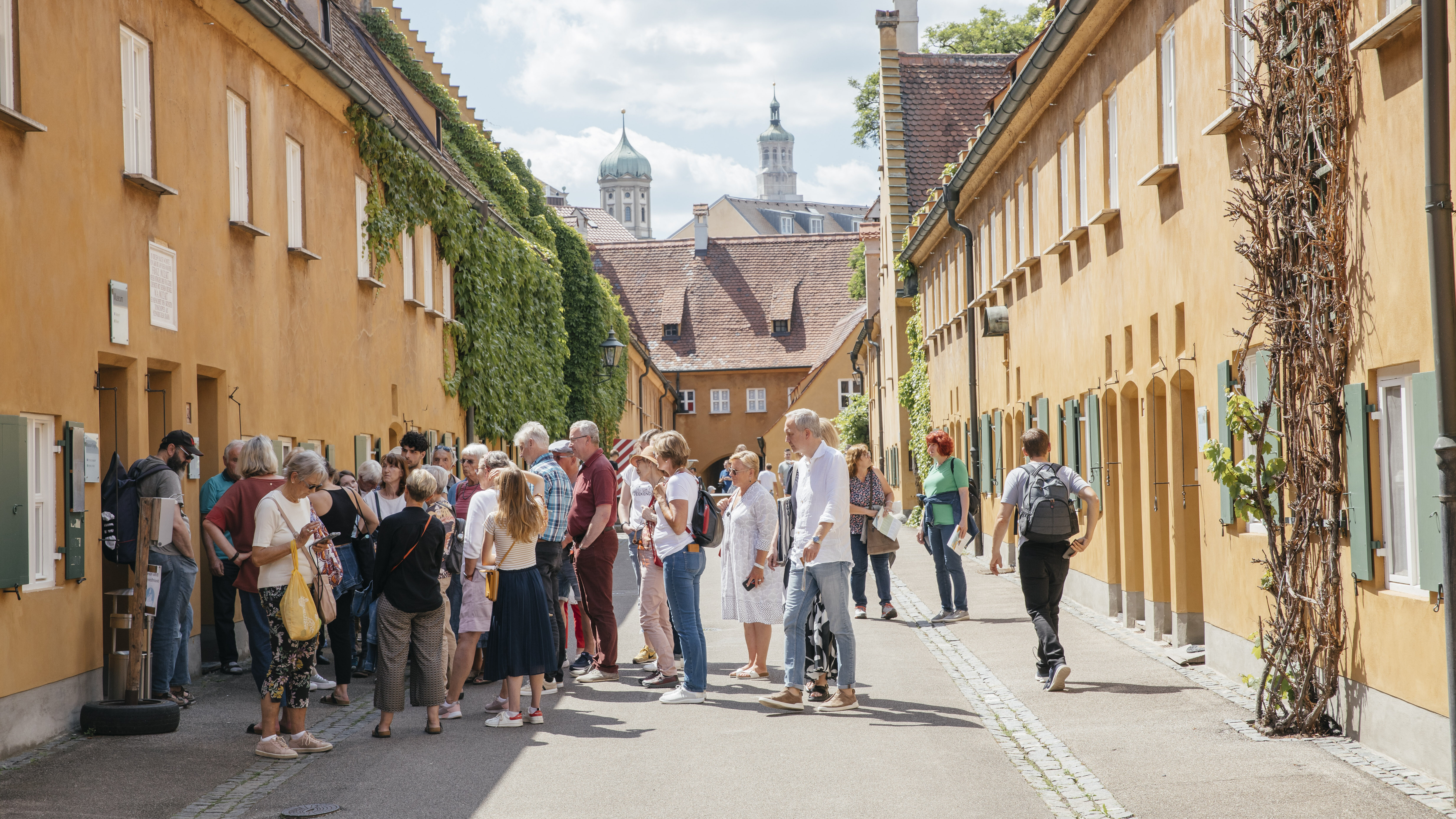 Zu sehen ist die Fuggerei. Auf den kleinen Strßaen tummelt sich einige Menschen, die an einer Führung durch die Fuggerei teilnehmen.