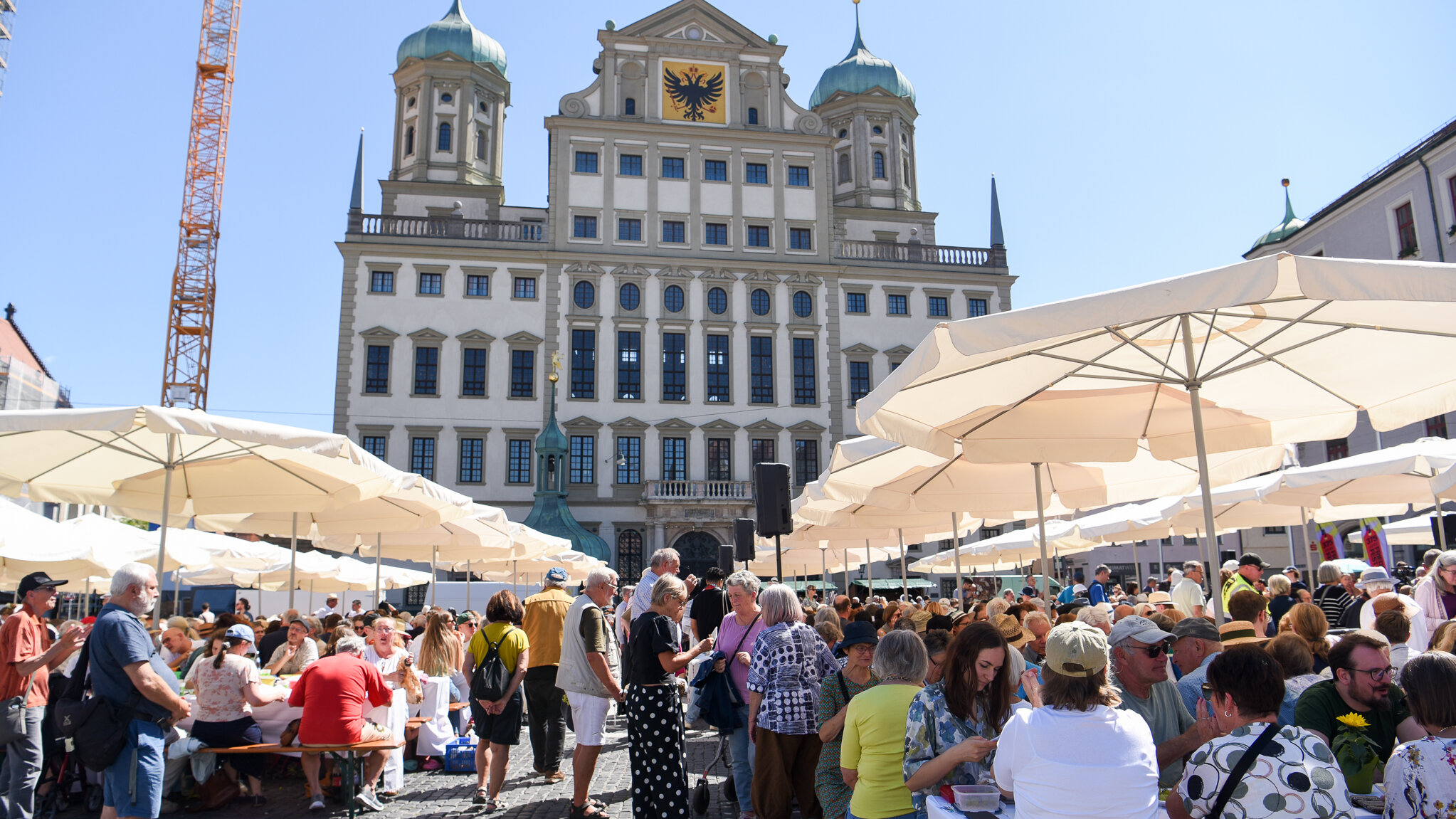 im Hintergrund das Augsburger Rathaus, im Vordergrund Menschen, die auf Bierbänken unter großen Sonnenschirmen sitzen.