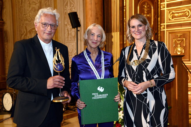 Ein Mann und zwei Frauen stehen im Goldenen Saal. Der Mann hält eine Skulptur in der Hand und die Frau in der Mitte eine Urkunde.