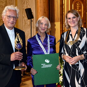 Ein Mann und zwei Frauen stehen im Goldenen Saal. Der Mann hält eine Skulptur in der Hand und die Frau in der Mitte eine Urkunde.