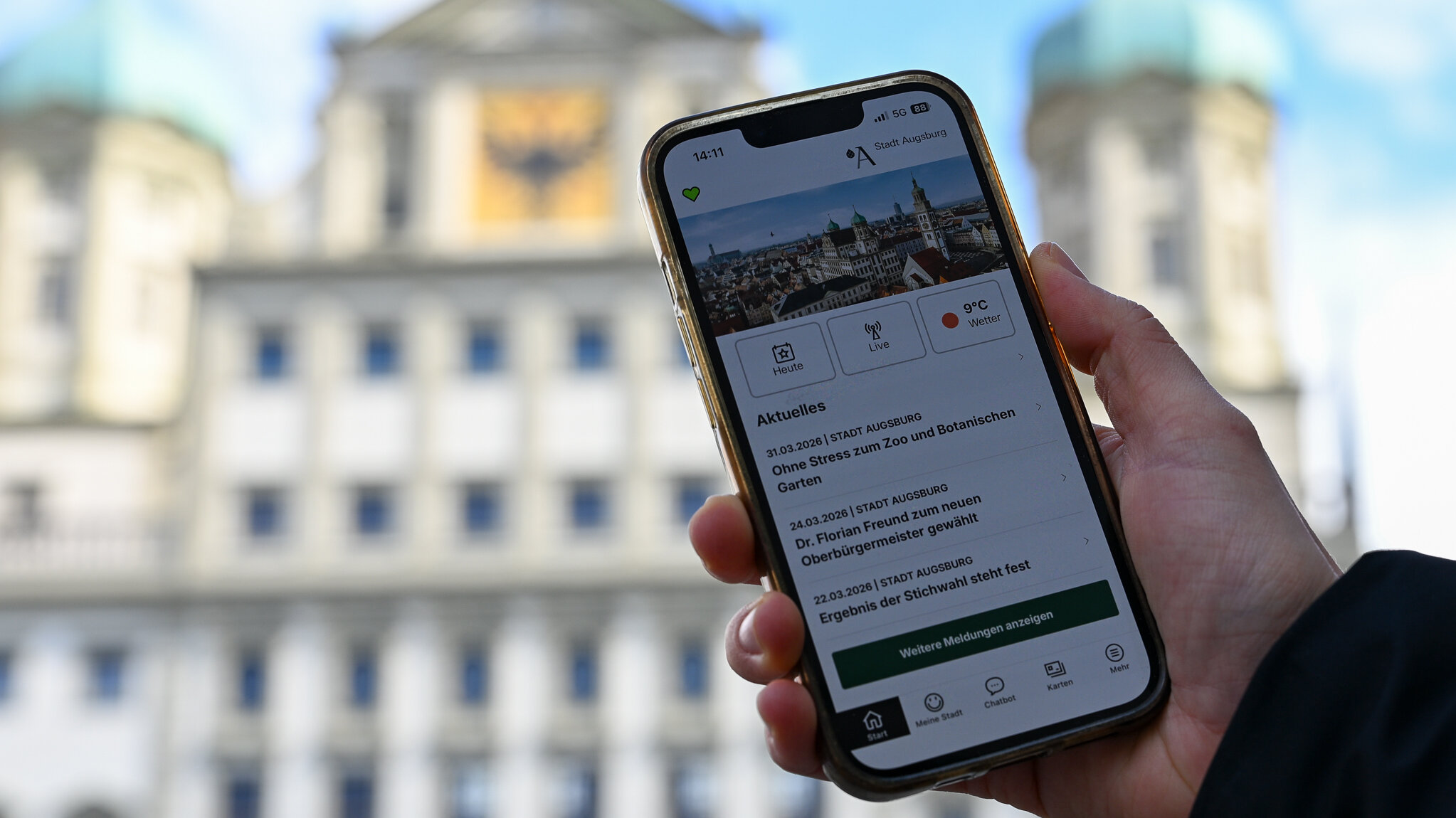 Zu sehen ist eine Hand, die ein Smartphone hält. Darauf ist die Augsburg App zu sehen. Im Hintergrund  das Augsburger Rathaus