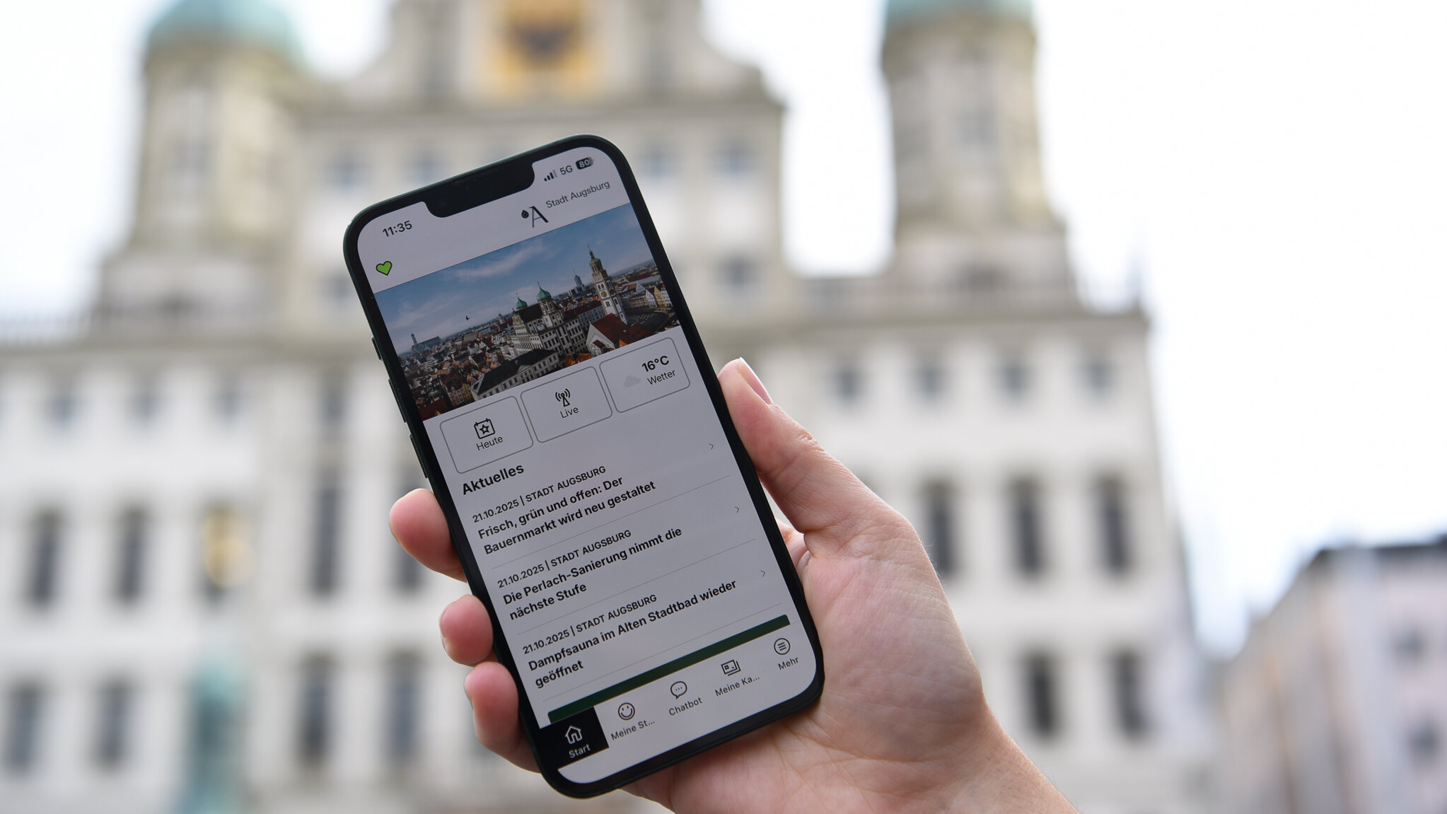 Smartphone zeigt die Augsburg App. Im Hintergrund ist das Augsburger Rathaus zu sehen